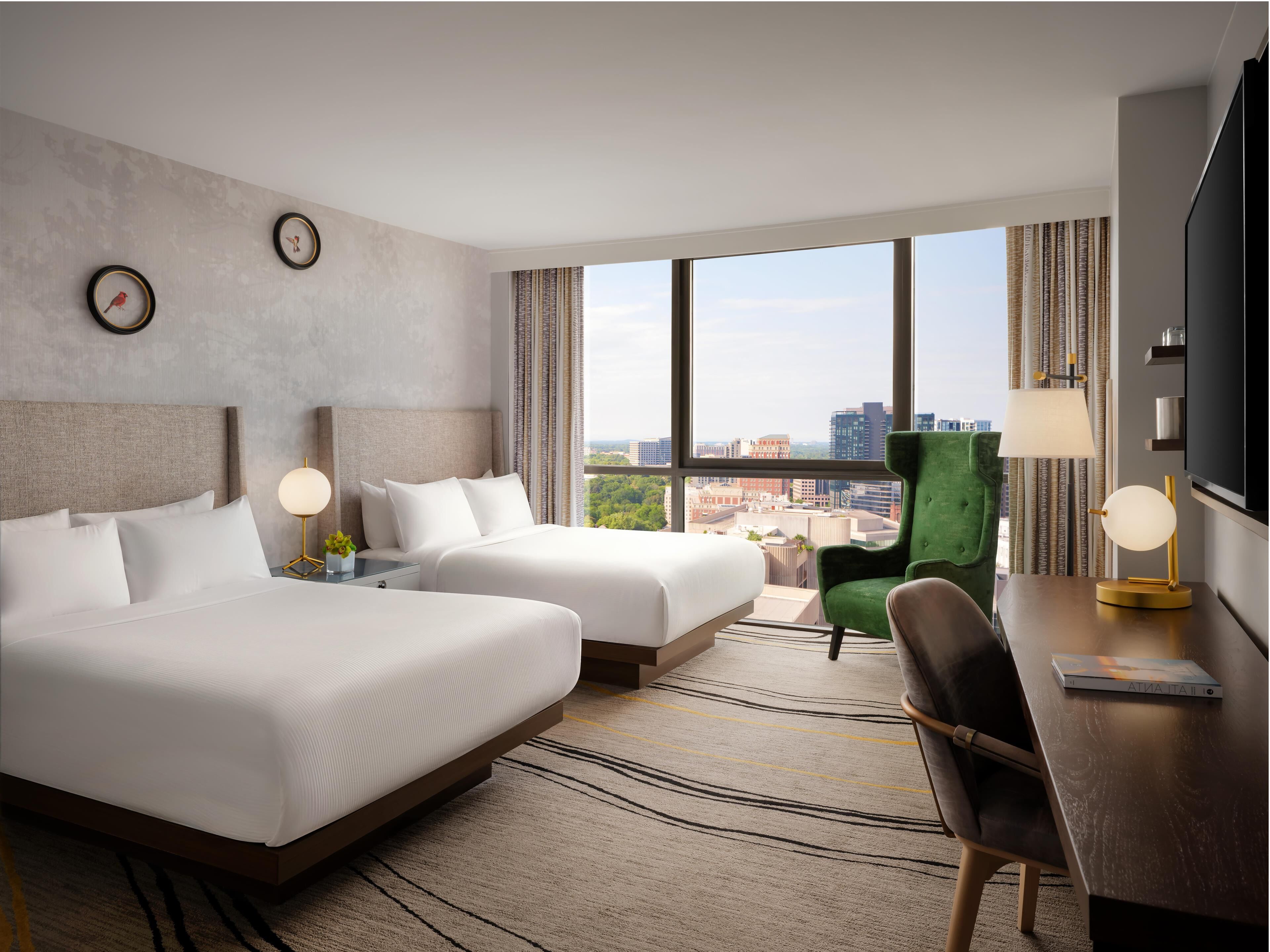 Hotelkamer met twee bedden, een groene fauteuil, een bureau met lamp, een televisie en grote ramen met uitzicht op de stad.