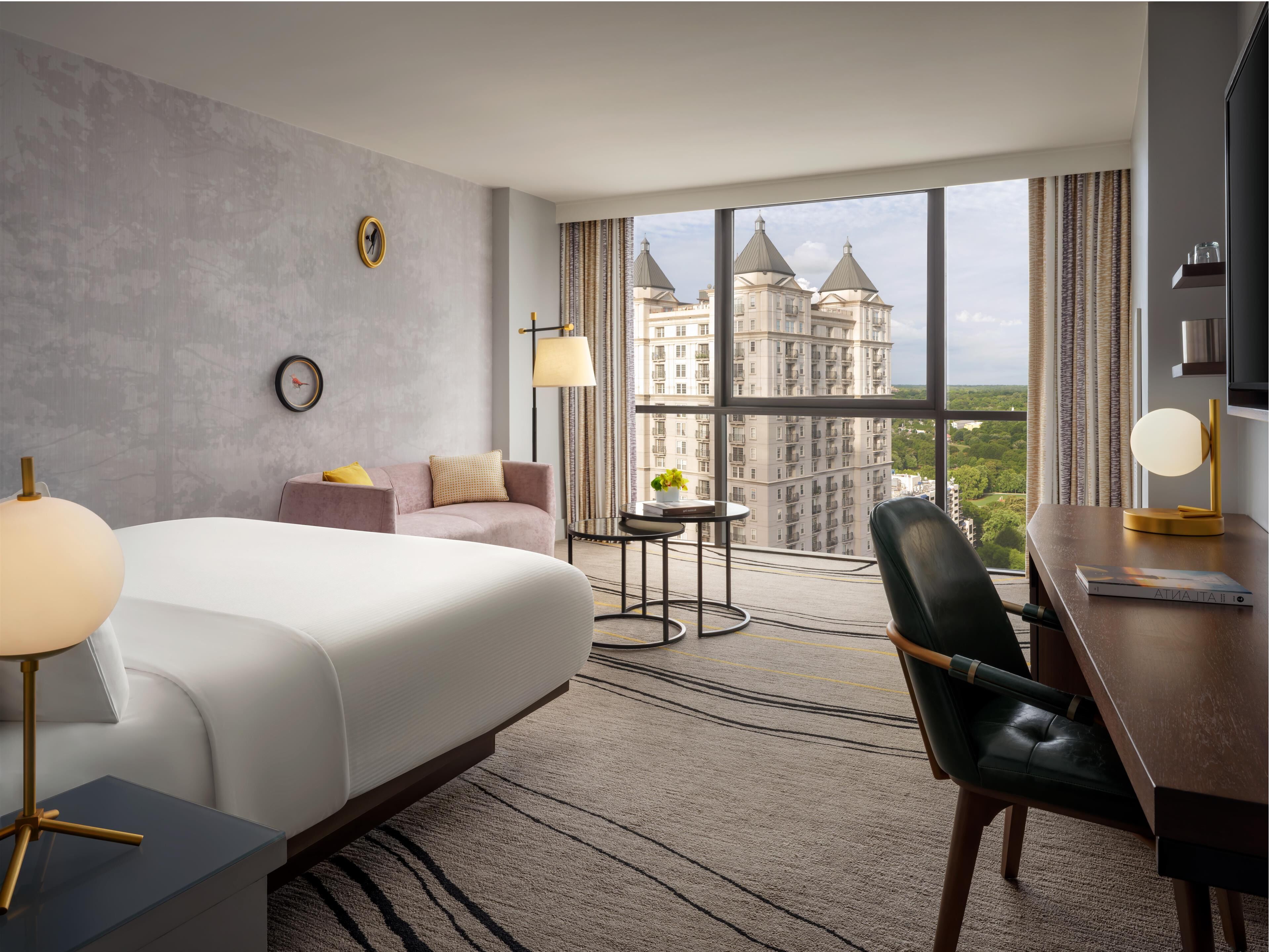 Une chambre d’hôtel avec un grand lit, un bureau avec une chaise, une petite table ronde et une vue sur un château, visible à travers de grandes fenêtres.