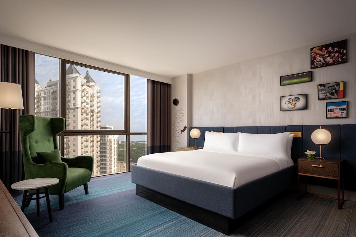 A hotel room with a large bed, a green armchair, a small round table, and a view of tall buildings.