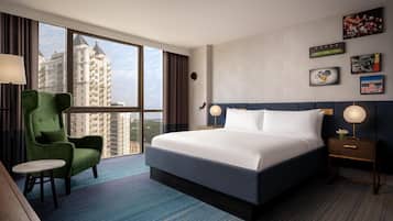 A hotel room with a large bed, a green armchair, a small round table, and a view of tall buildings through the window.