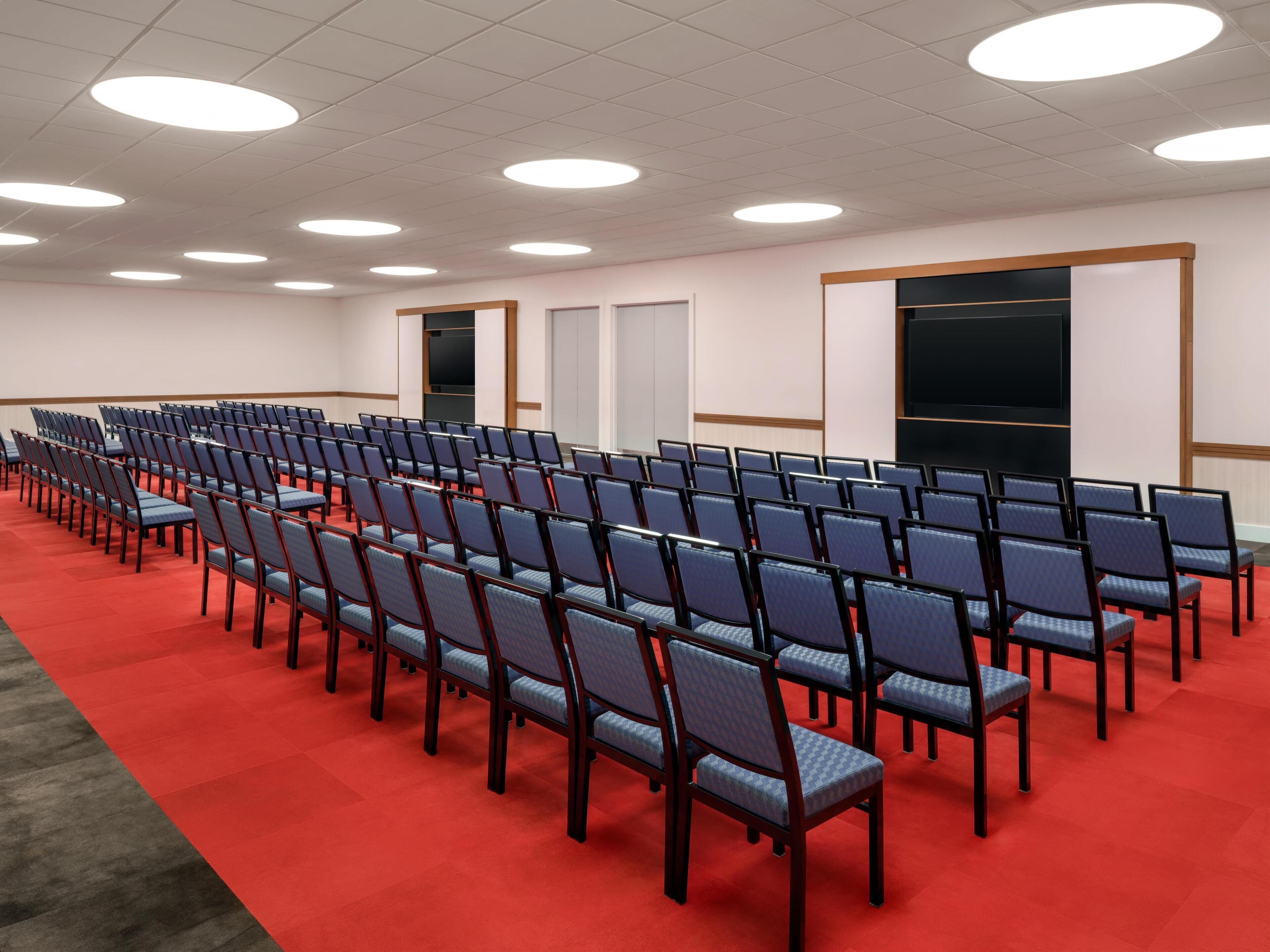 Ein Konferenzraum mit Stuhlreihen, einem roten Teppich und Deckenleuchten.