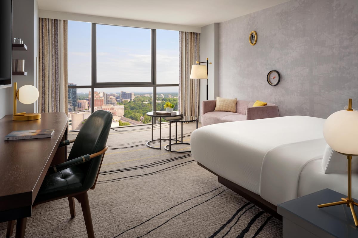 A hotel room with a large window, a bed, a desk, a chair, a small table, and a lamp.