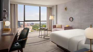 A hotel room with a large window, a bed, a desk, a chair, a small table, and a lamp.