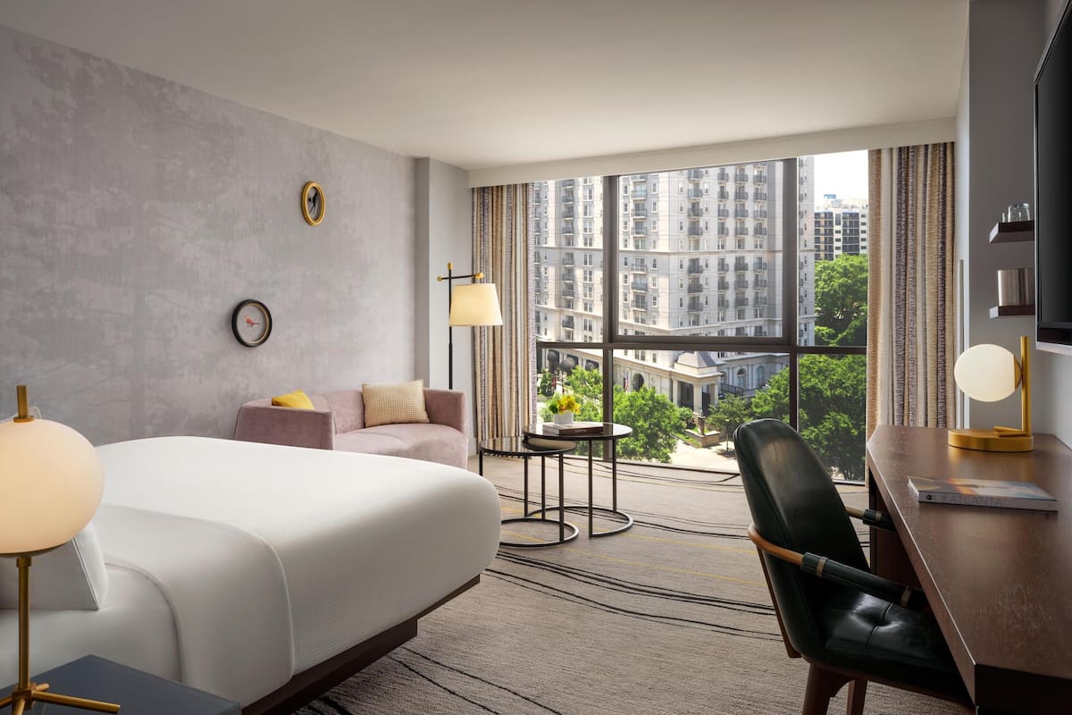 A hotel room with a large bed, a desk with a chair, a small table with a lamp, a sofa, and a view of buildings through large windows.