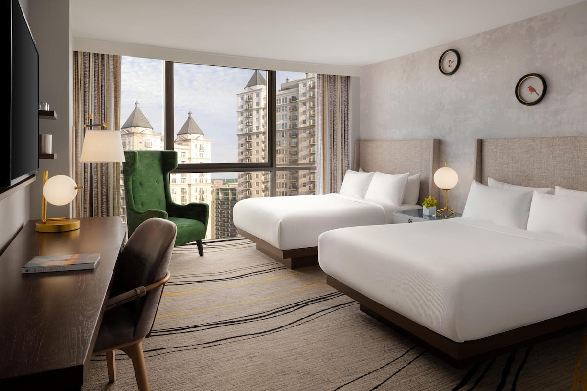 A hotel room with two beds, a desk, a chair, a TV, and a view of tall buildings.