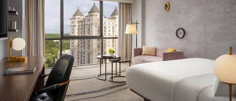 A hotel room with a large bed, a desk with a chair, a small table with a lamp, and a view of a castle through large windows.