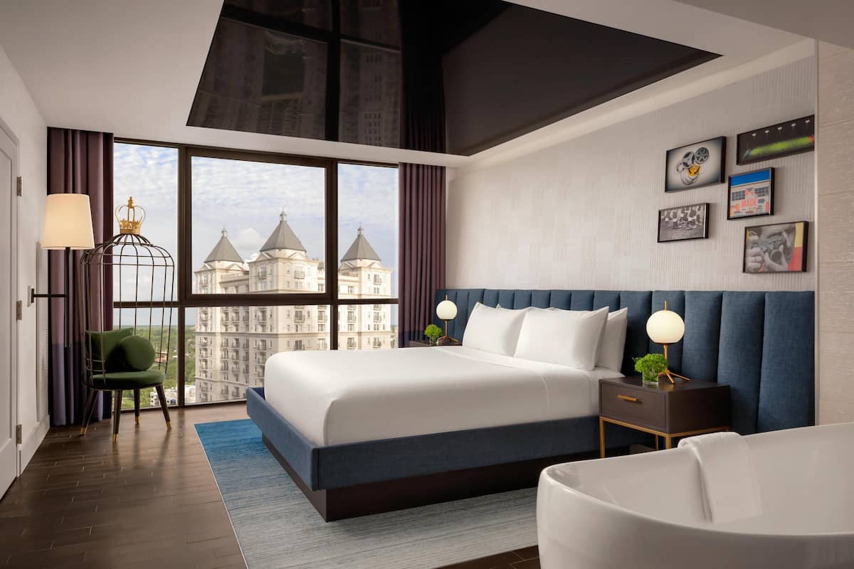 A modern hotel bedroom with a large bed, a view of city buildings, and a blue headboard.