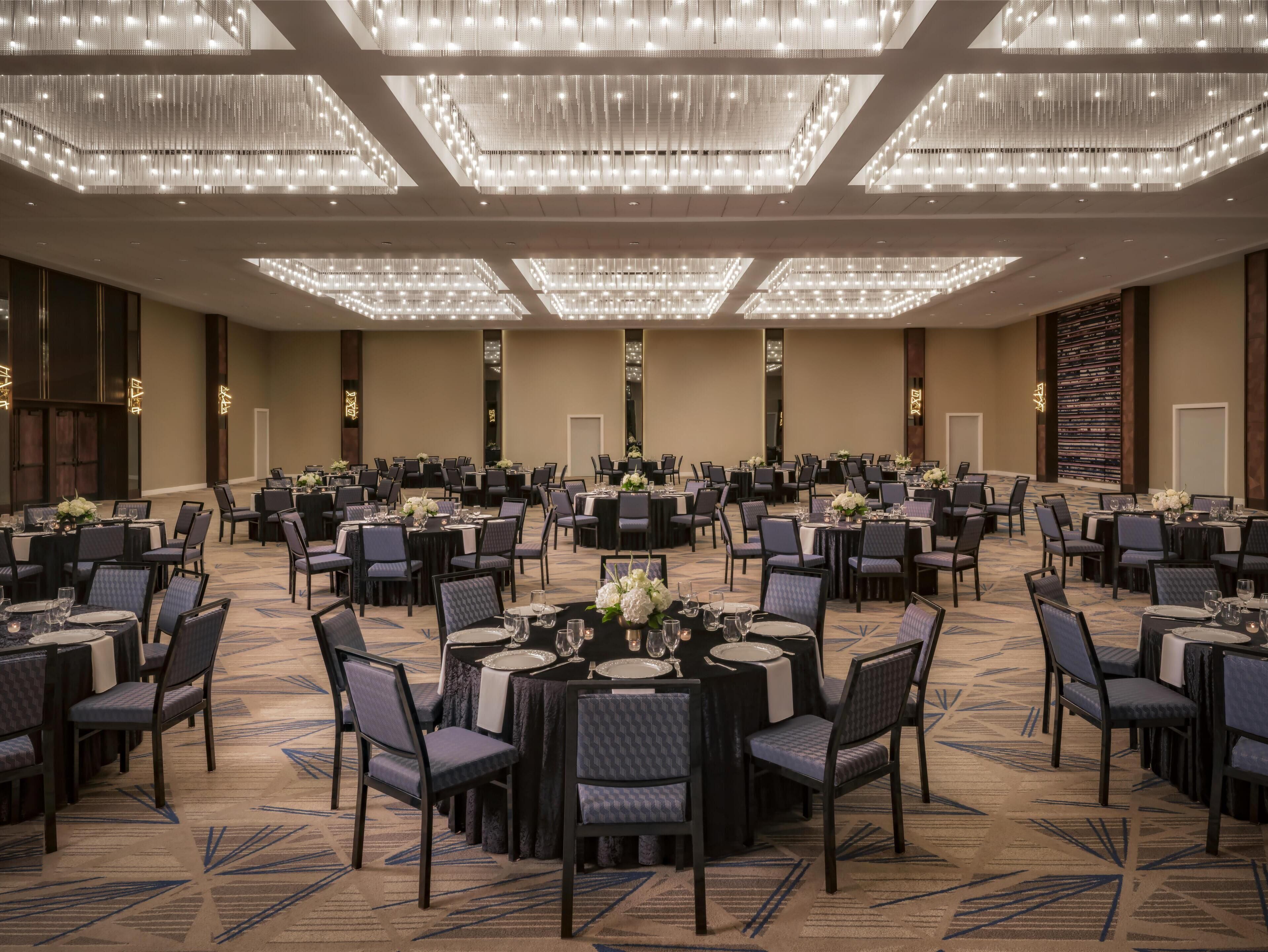 A large banquet hall with round tables, chairs, and chandeliers.