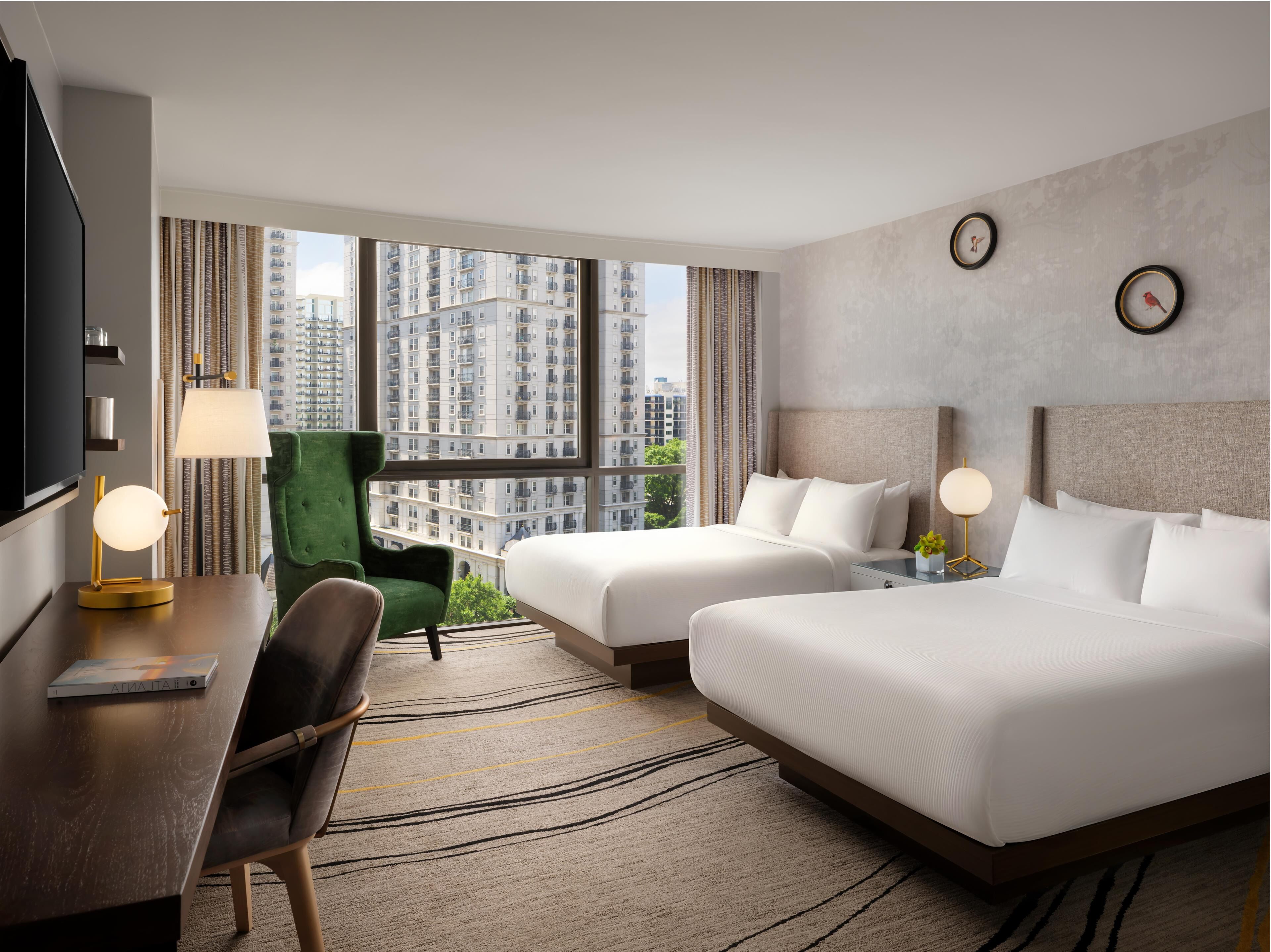 A hotel room with two beds, a desk, a chair, a TV, and a large window with a city view.