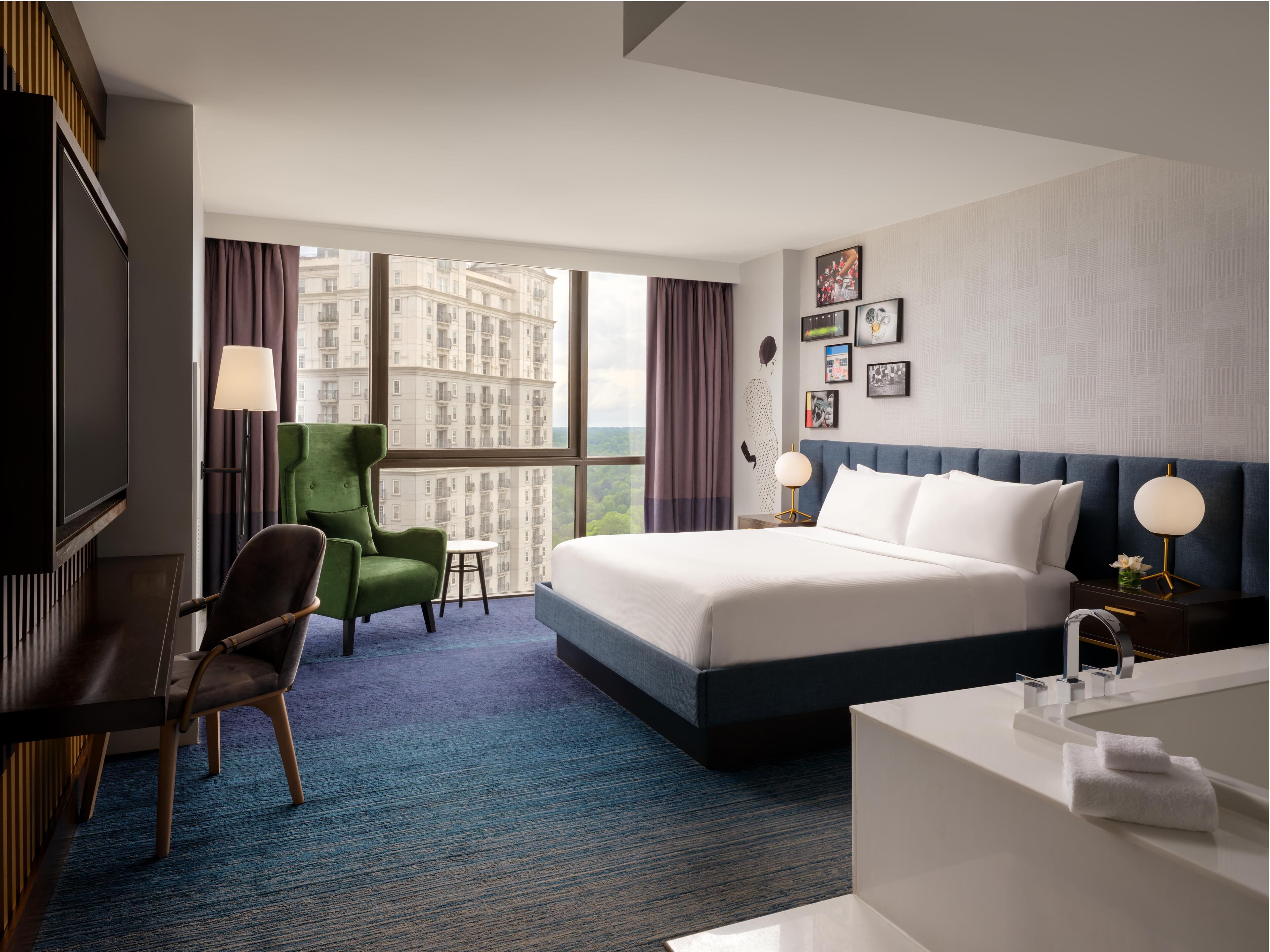 A hotel room with a large bed, a desk, a chair, a small table, a nightstand, a lamp, and a view of a cityscape through large windows.