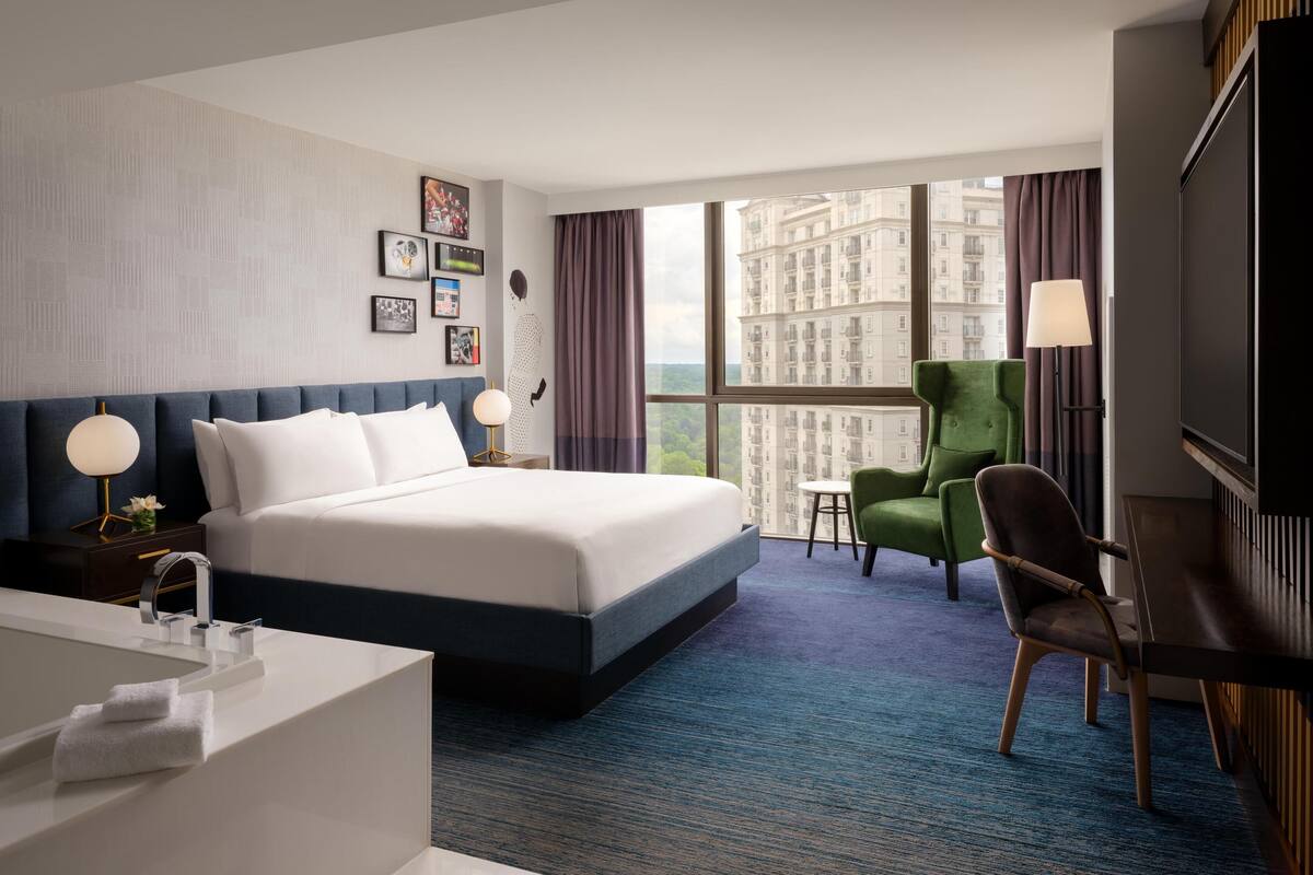 A modern hotel room with a large bed, a desk, a green armchair, and a view of a cityscape through large windows.