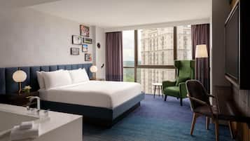 Een moderne hotelkamer met een groot bed, een bureau, een groene fauteuil en een stadsgezicht door grote ramen.