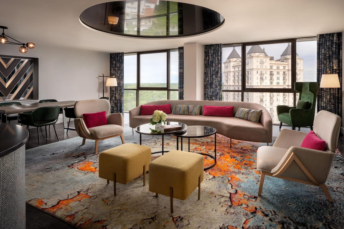 A modern hotel room with a large window, a round coffee table, a sofa, and two armchairs.