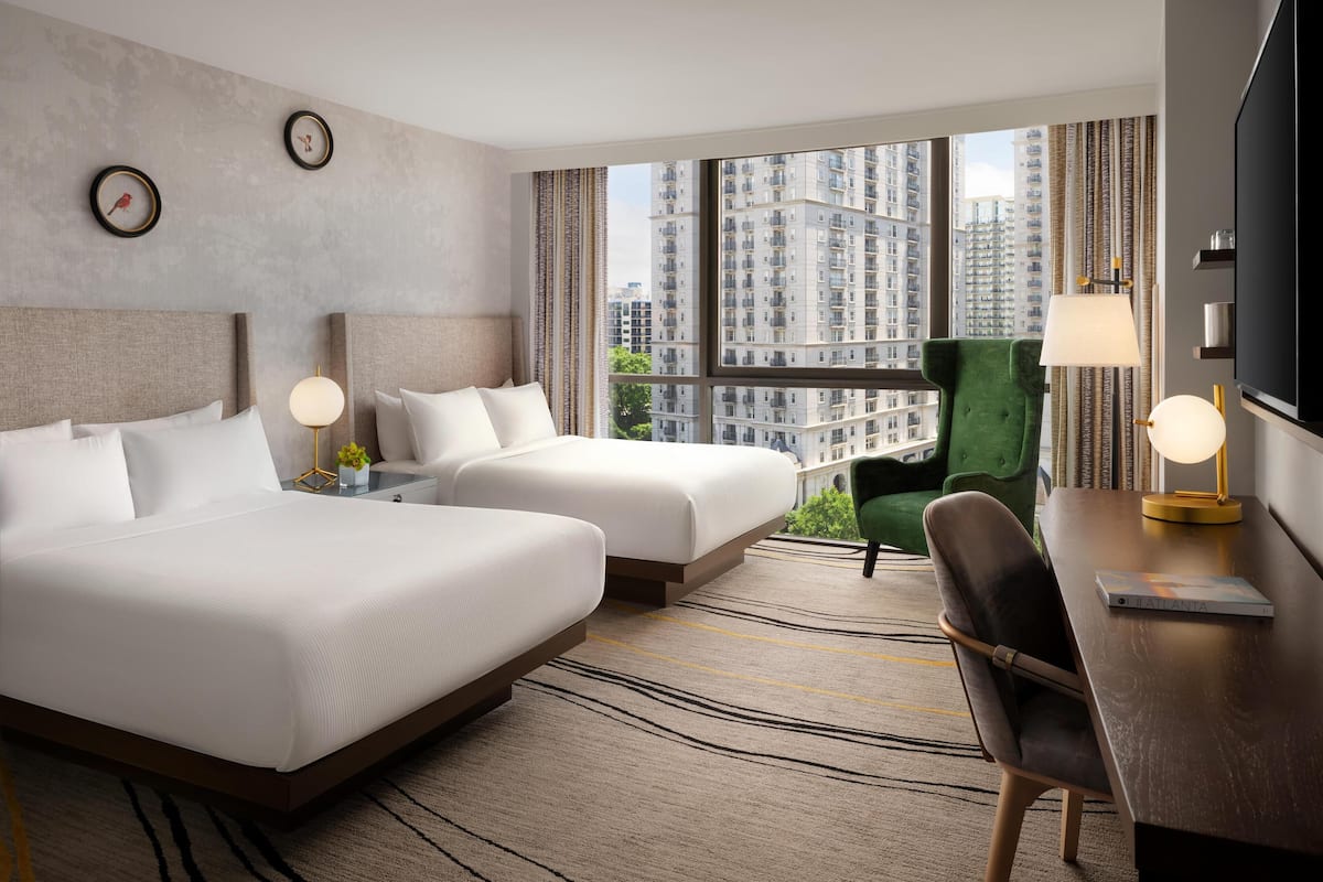 A hotel room with two beds, a green armchair, a TV, and a view of high-rise buildings.