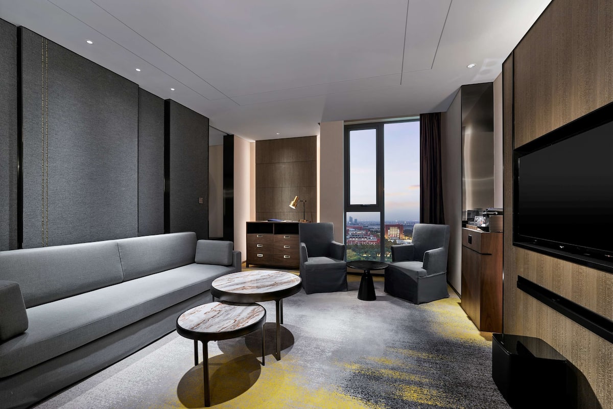 A modern hotel room with a sofa, two armchairs, a coffee table, a TV, and a city view through large windows.