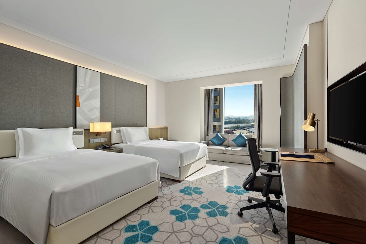 A hotel room with two beds, a desk, a chair, a TV, and a large window with a city view.