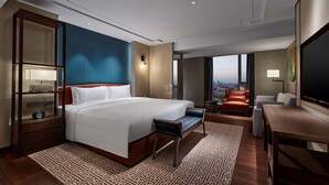 Une chambre d’hôtel moderne dotée d’un grand lit, d’un coin salon avec un canapé et des fauteuils, d’un bureau et d’une grande fenêtre donnant sur la ville.