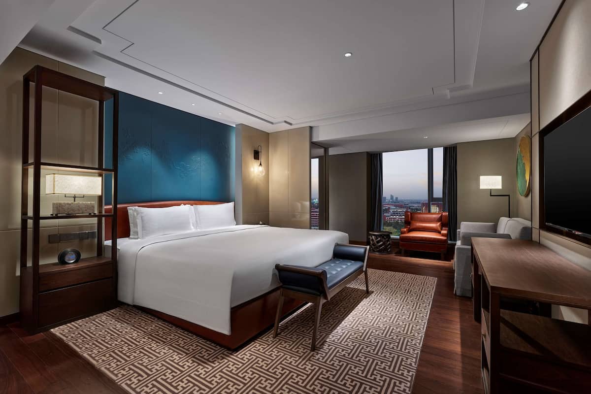 A modern hotel room with a large bed, a seating area with a sofa and armchairs, a desk, and a large window offering a city view.