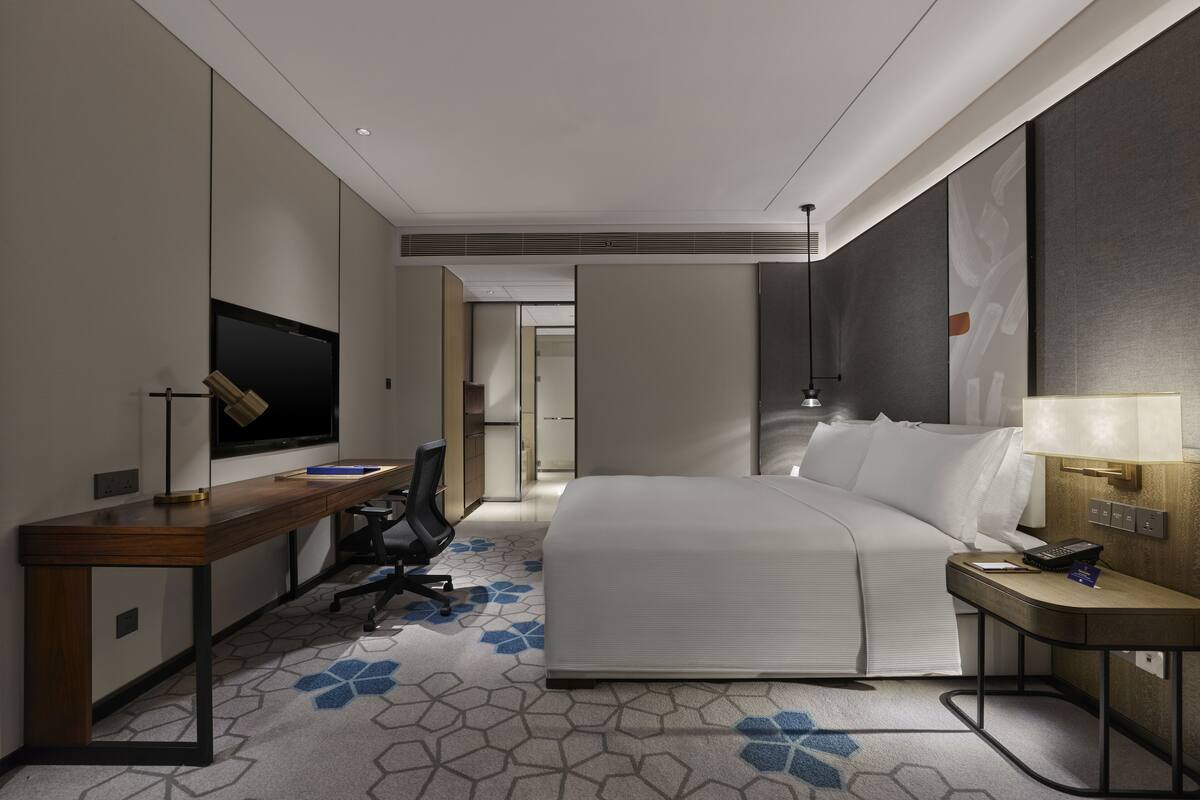A hotel room with a bed, desk, chair, TV, and a lamp.