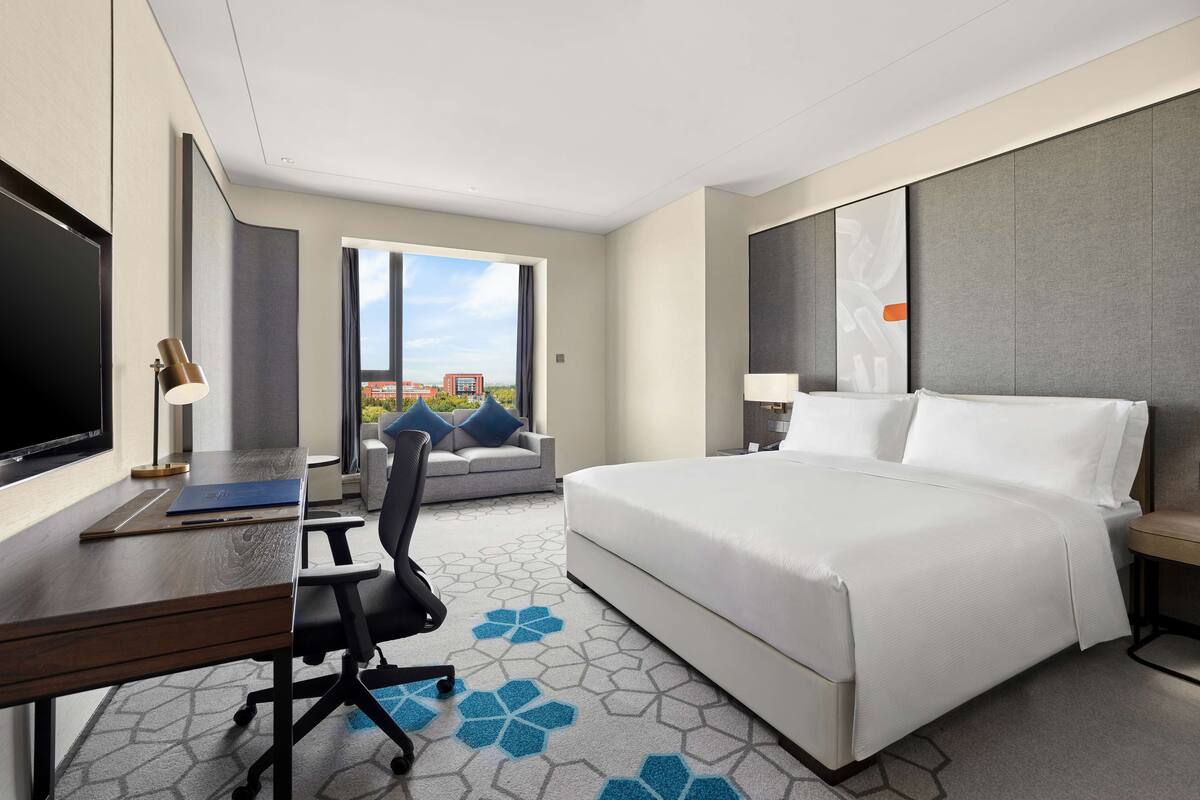 A hotel room with a bed, desk, chair, TV, and a view of the outdoors.