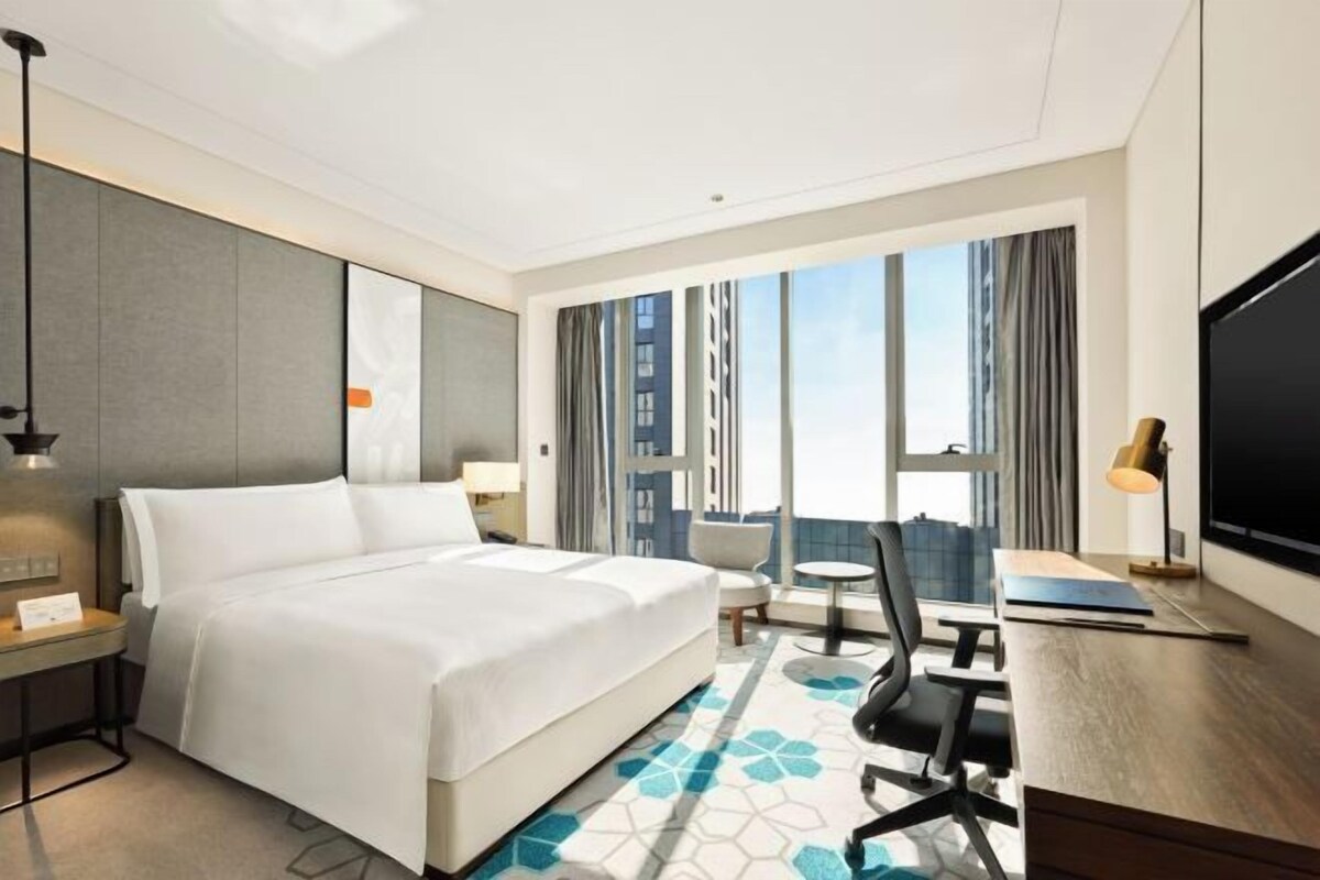 A hotel room with a large bed, a desk, a chair, a TV, and a view of the city.