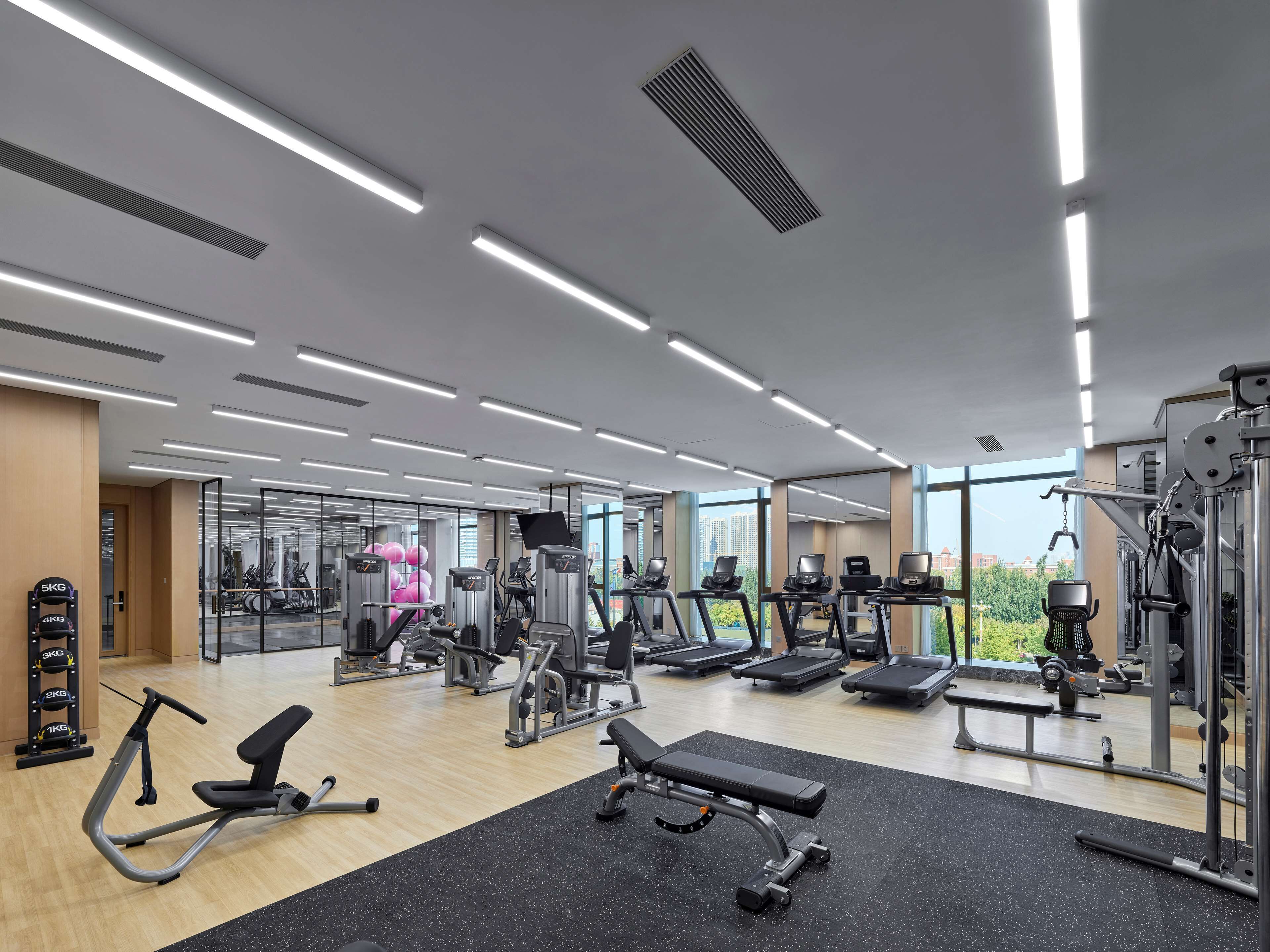 A modern gym with various exercise equipment, large windows, and a view of the city.