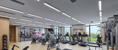 A modern gym with various exercise equipment, large windows, and a view of the city.