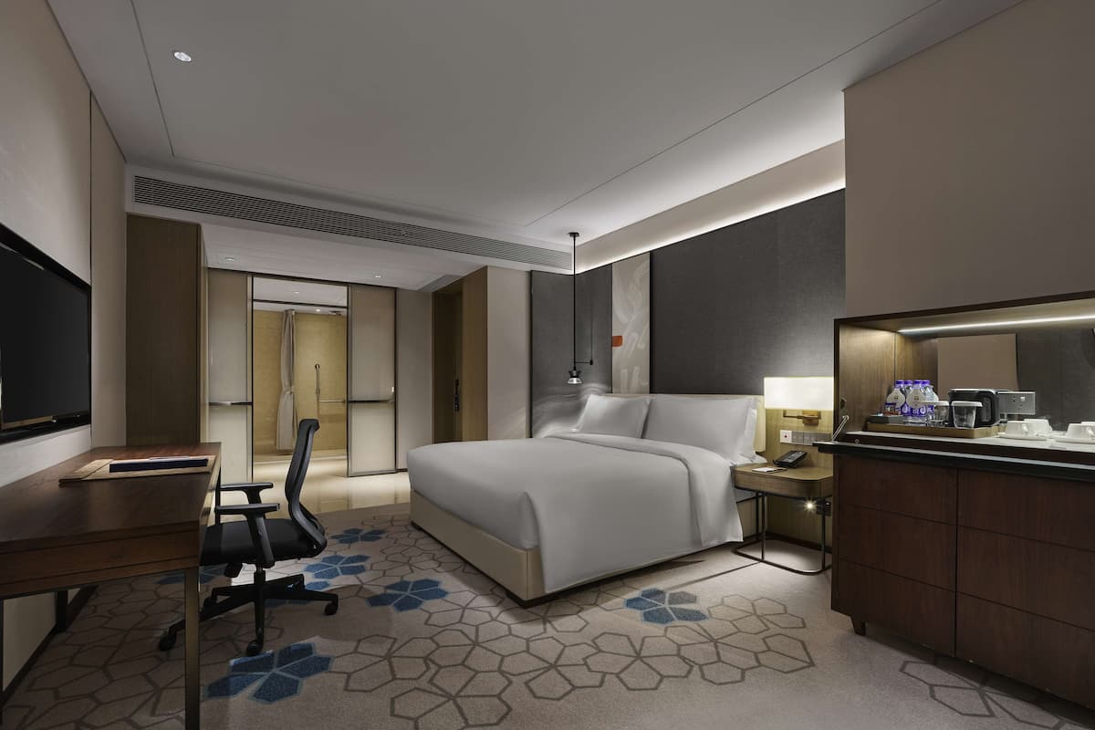 A modern hotel room with a large bed, a desk, a chair, a TV, and a bathroom visible through an open door.