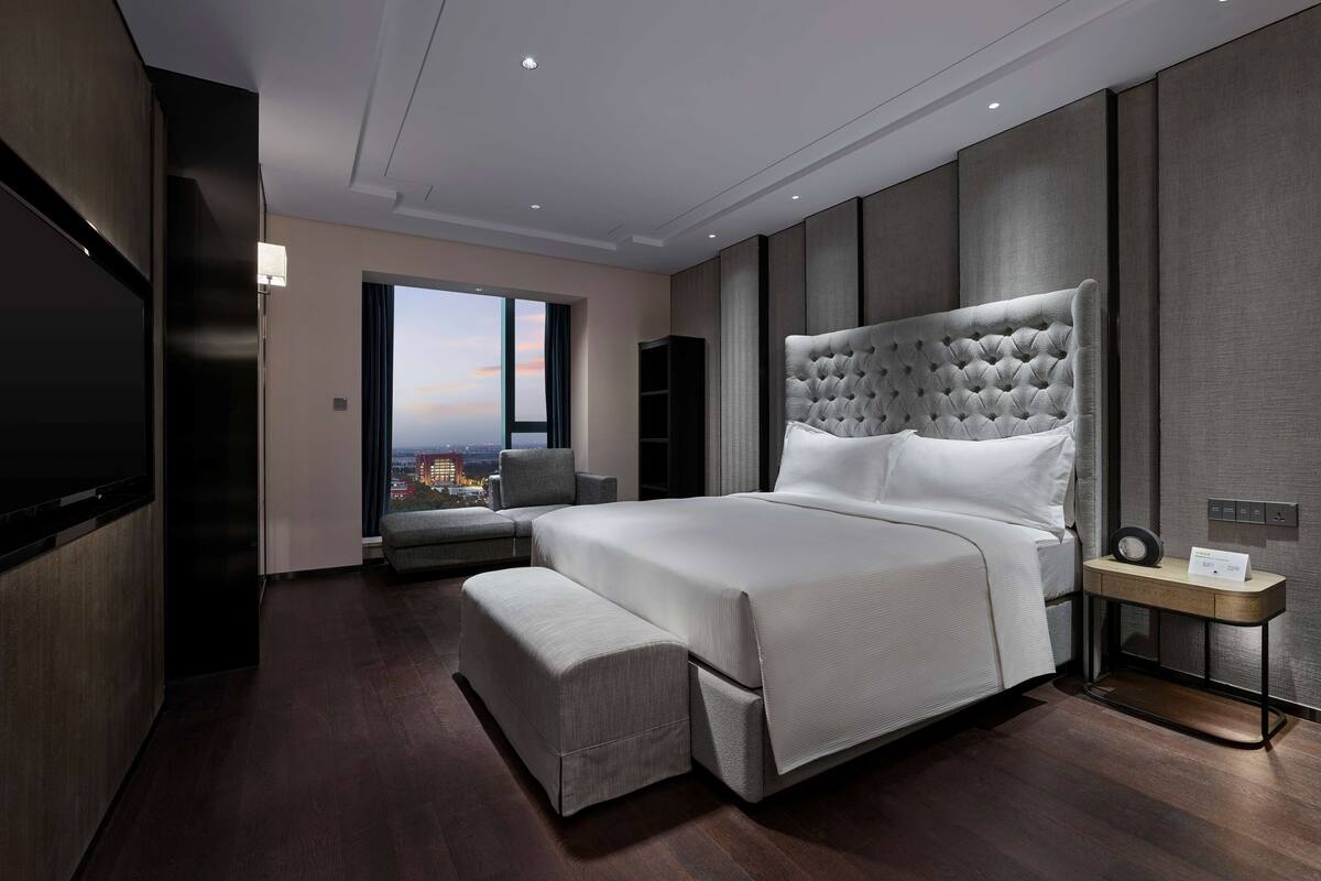 A modern hotel room with a large bed, a seating area, and a view of the cityscape.