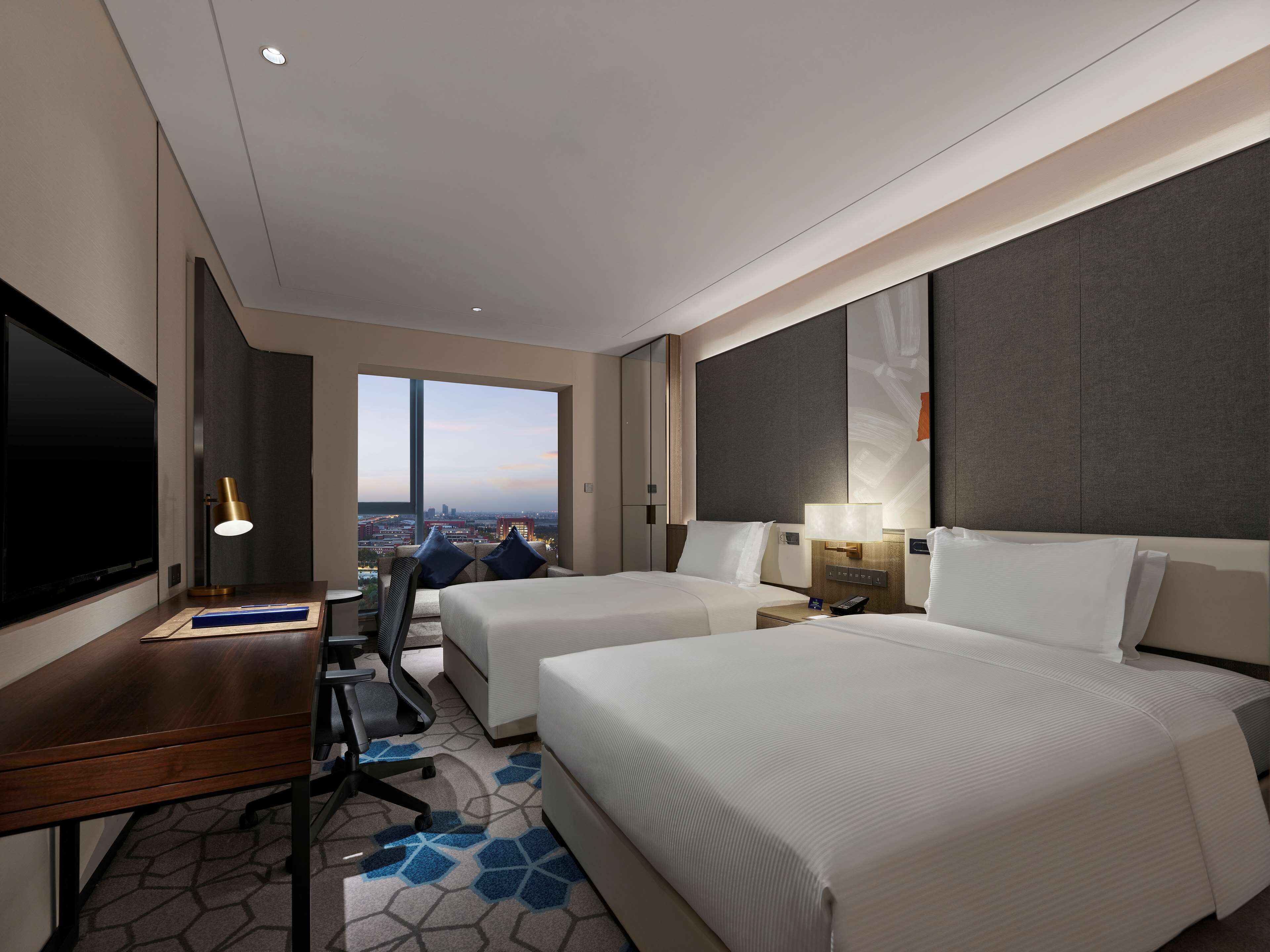 Une chambre d’hôtel avec deux lits, un bureau, une télévision et une grande fenêtre donnant sur la ville.