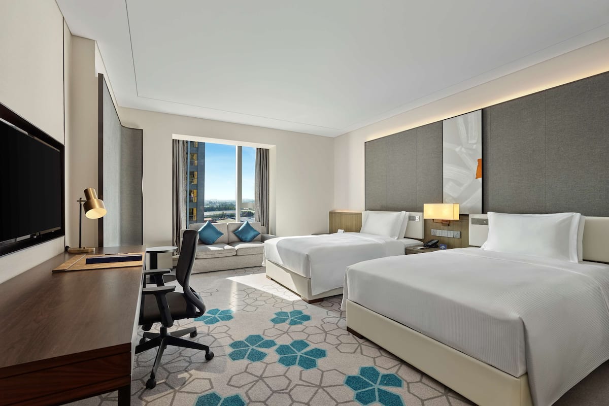 A hotel room with two beds, a desk, a TV, and a large window with a city view.