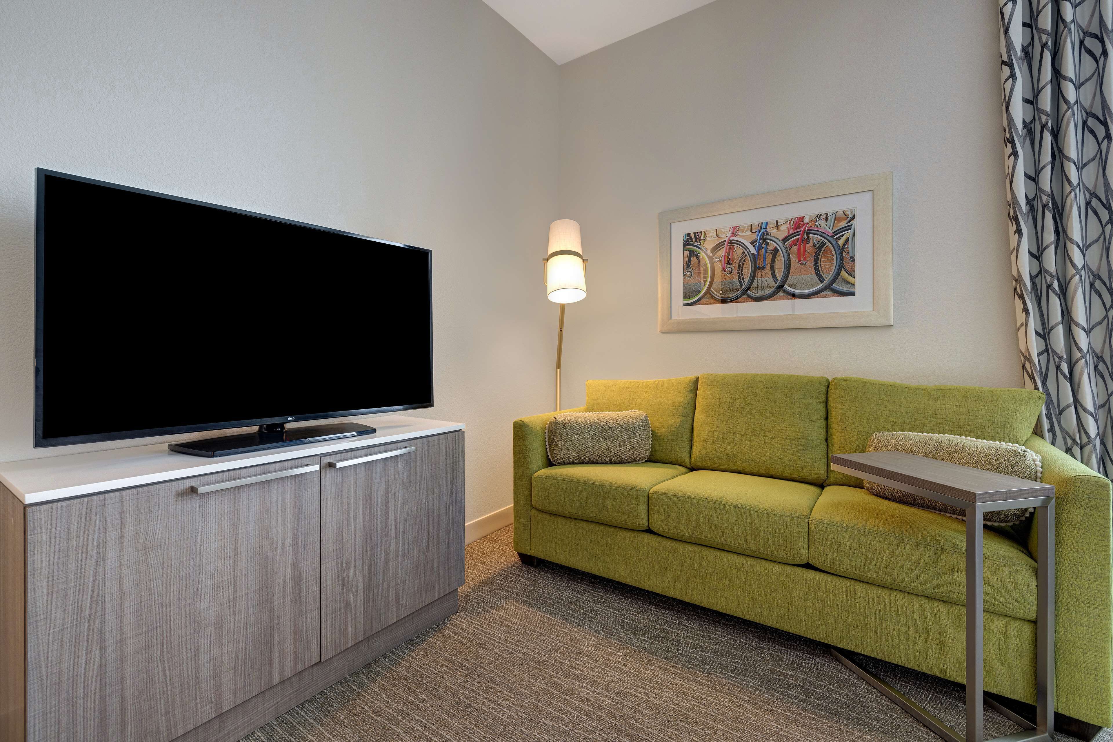 Una sala moderna con un televisor de pantalla plana sobre un mueble de madera, un sofá verde, una pequeña mesita auxiliar y una lámpara.