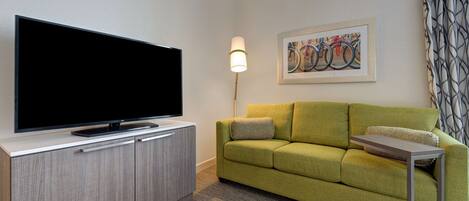 Una sala moderna con un televisor de pantalla plana sobre un mueble de madera, un sofá verde, una pequeña mesita auxiliar y una lámpara.