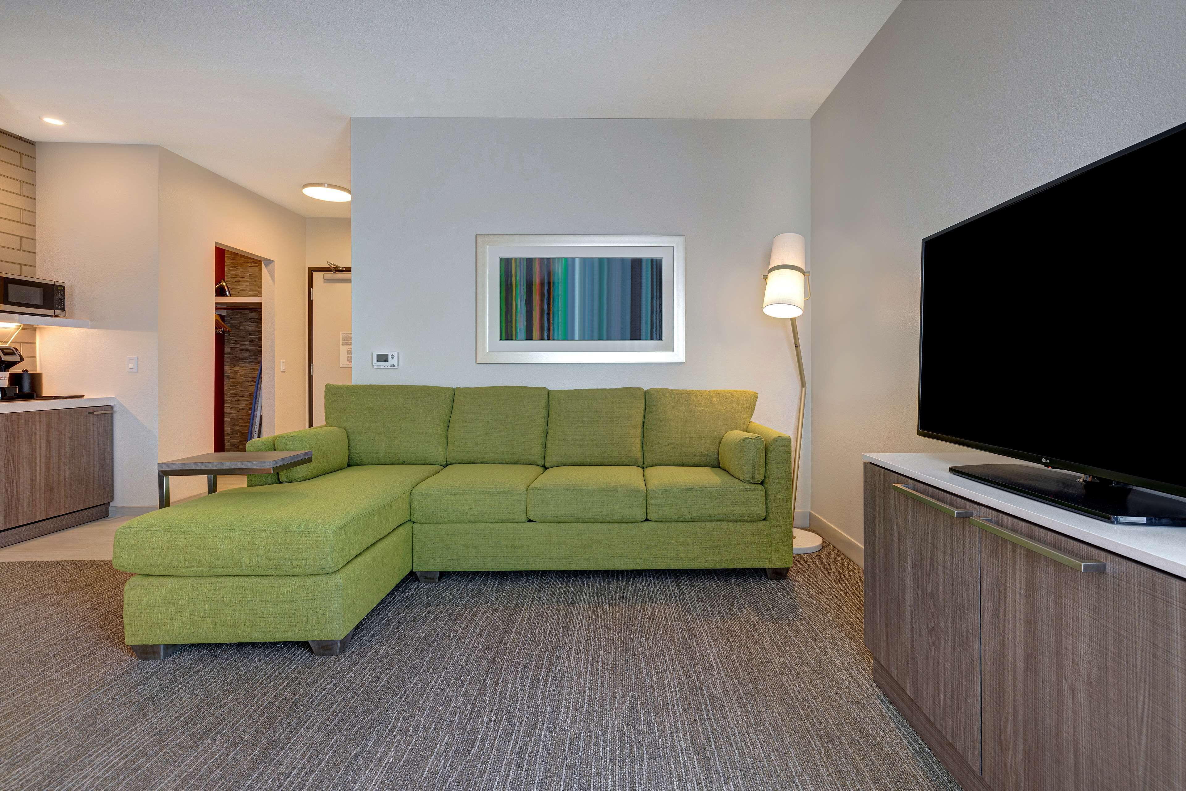 A modern living room with a green sectional sofa, a flat-screen TV, a framed abstract painting, and a small table.