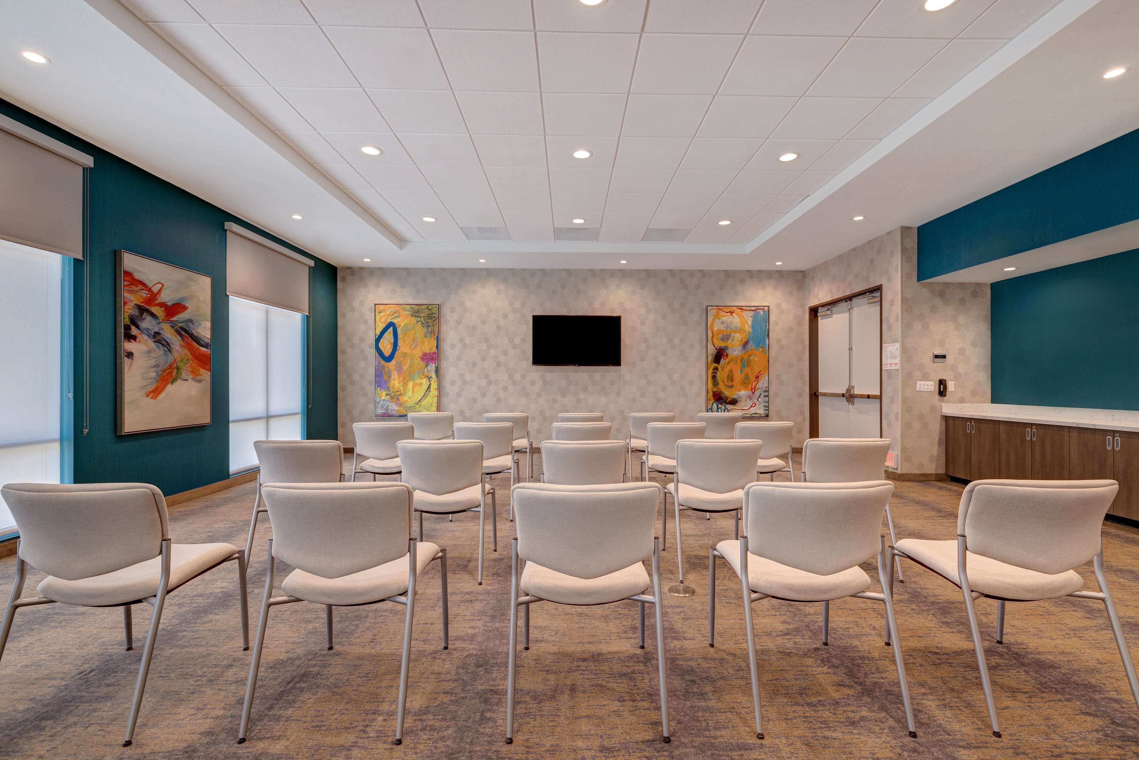 A conference room with rows of white chairs, a flat-screen TV on the wall, and abstract paintings on the walls.