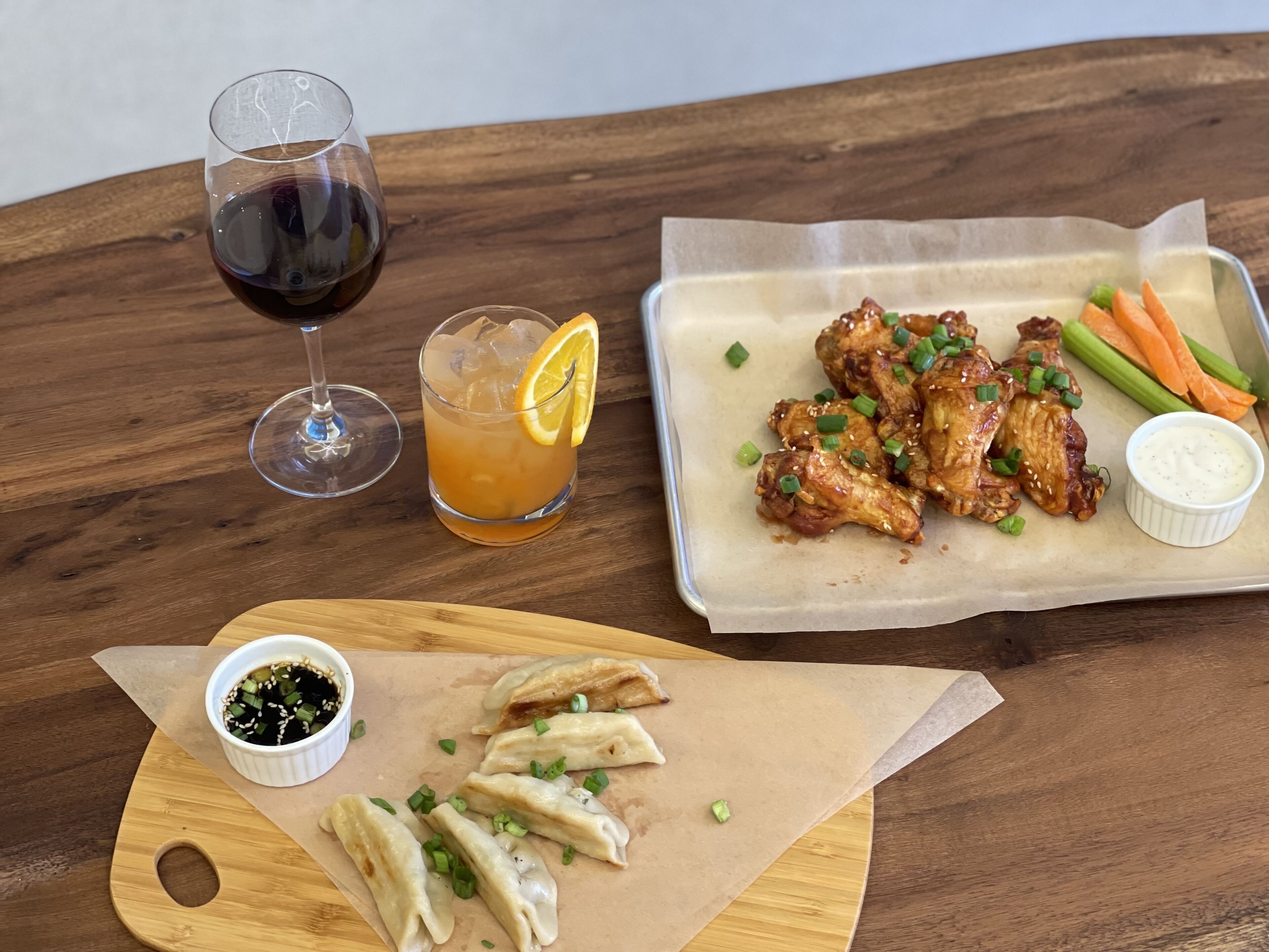 A wooden table with a glass of red wine, a glass of iced tea with a lemon slice, a plate of chicken wings with vegetables and dipping sauce, and a wooden board with dumplings and dipping sauce.