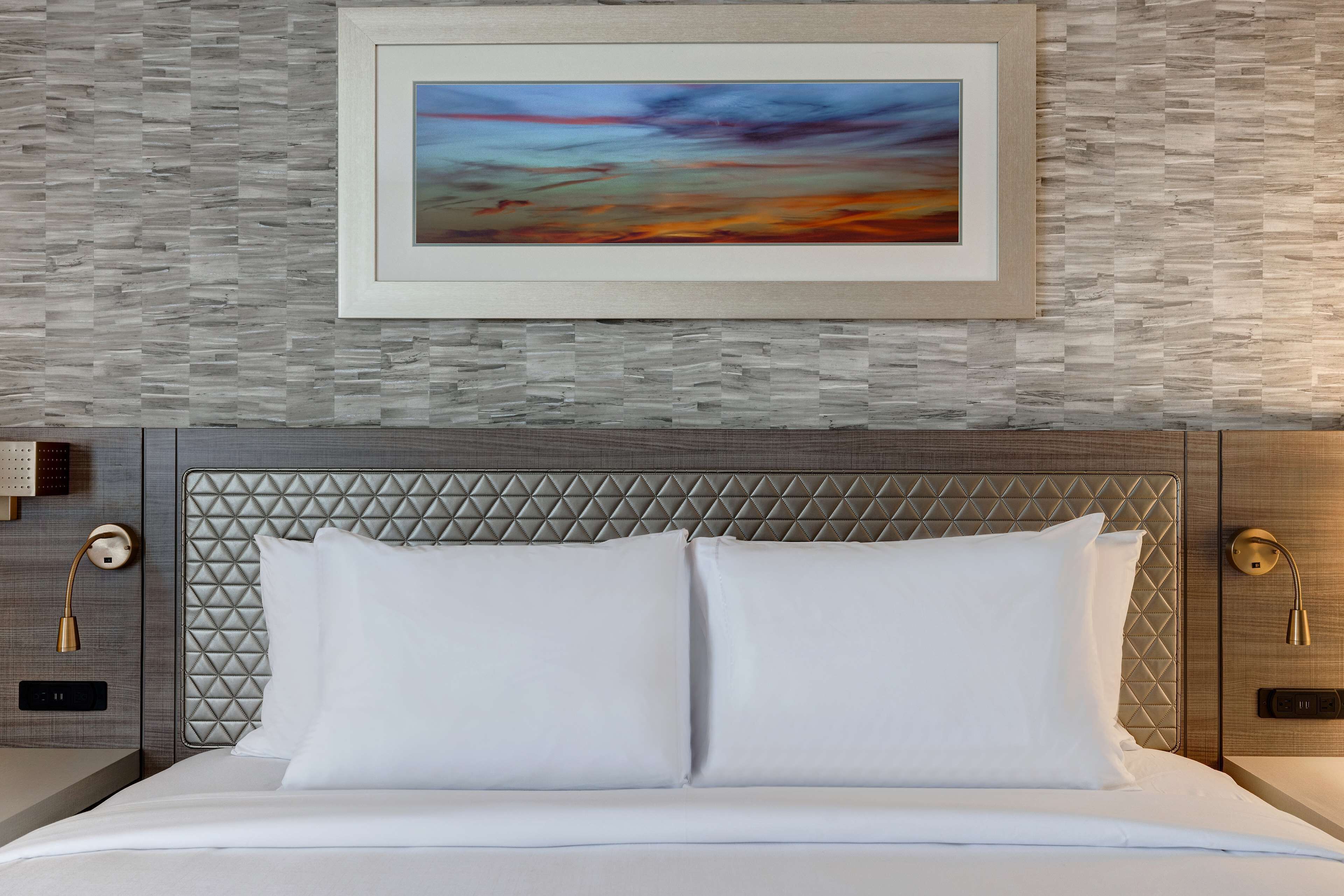 Un lit bien fait, avec du linge de lit blanc, un mur à la texture intéressante orné d’un tableau abstrait encadré, et une tête de lit moderne avec un motif géométrique.