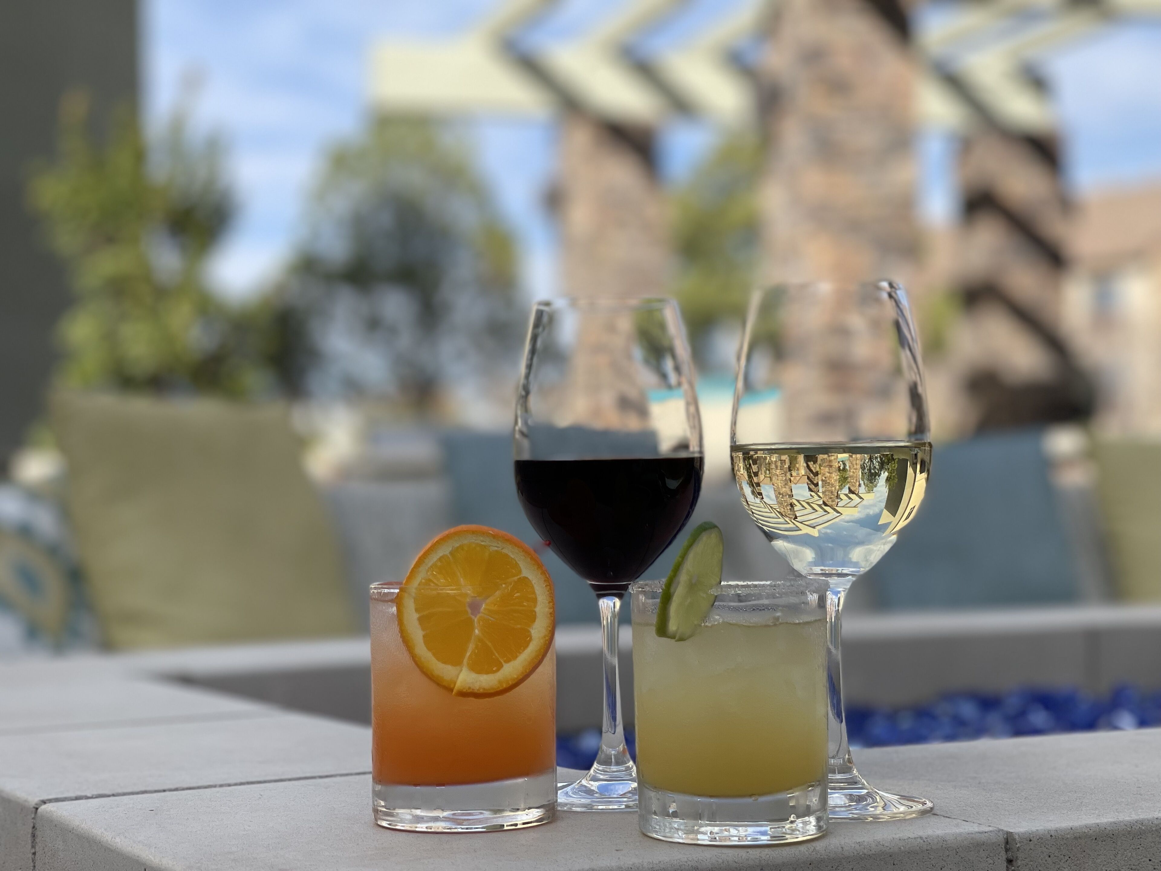 Three glasses of beverages on a terrace with a pool and a building in the background.