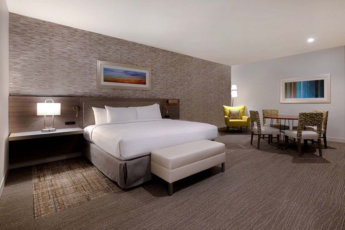 A hotel room with a large bed, a bedside table with a lamp, a small table with chairs, and a painting on the wall.