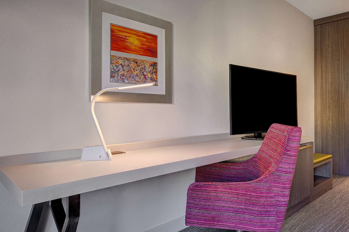 A modern hotel room with a white desk, a flat-screen TV, a colorful chair, and a framed artwork on the wall.