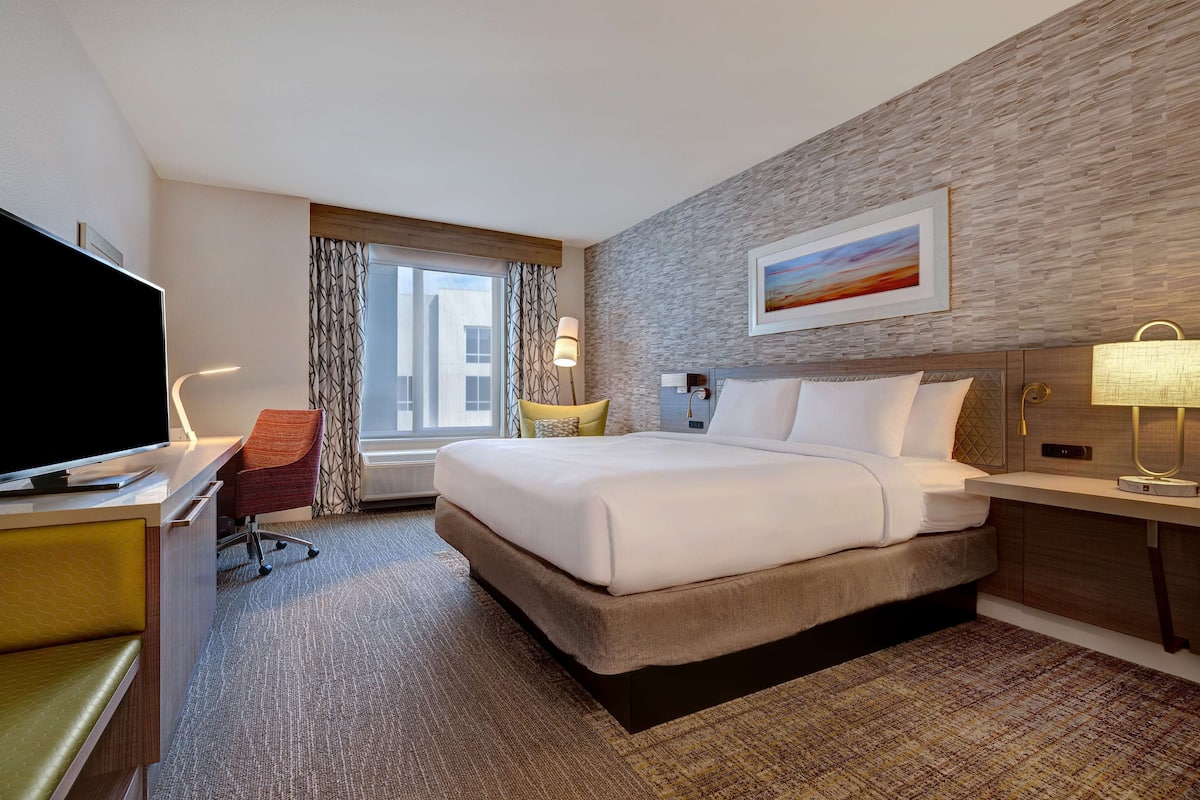 A hotel room with a large bed, a desk with a flat-screen TV, a chair, a window with curtains, and a painting on the wall.