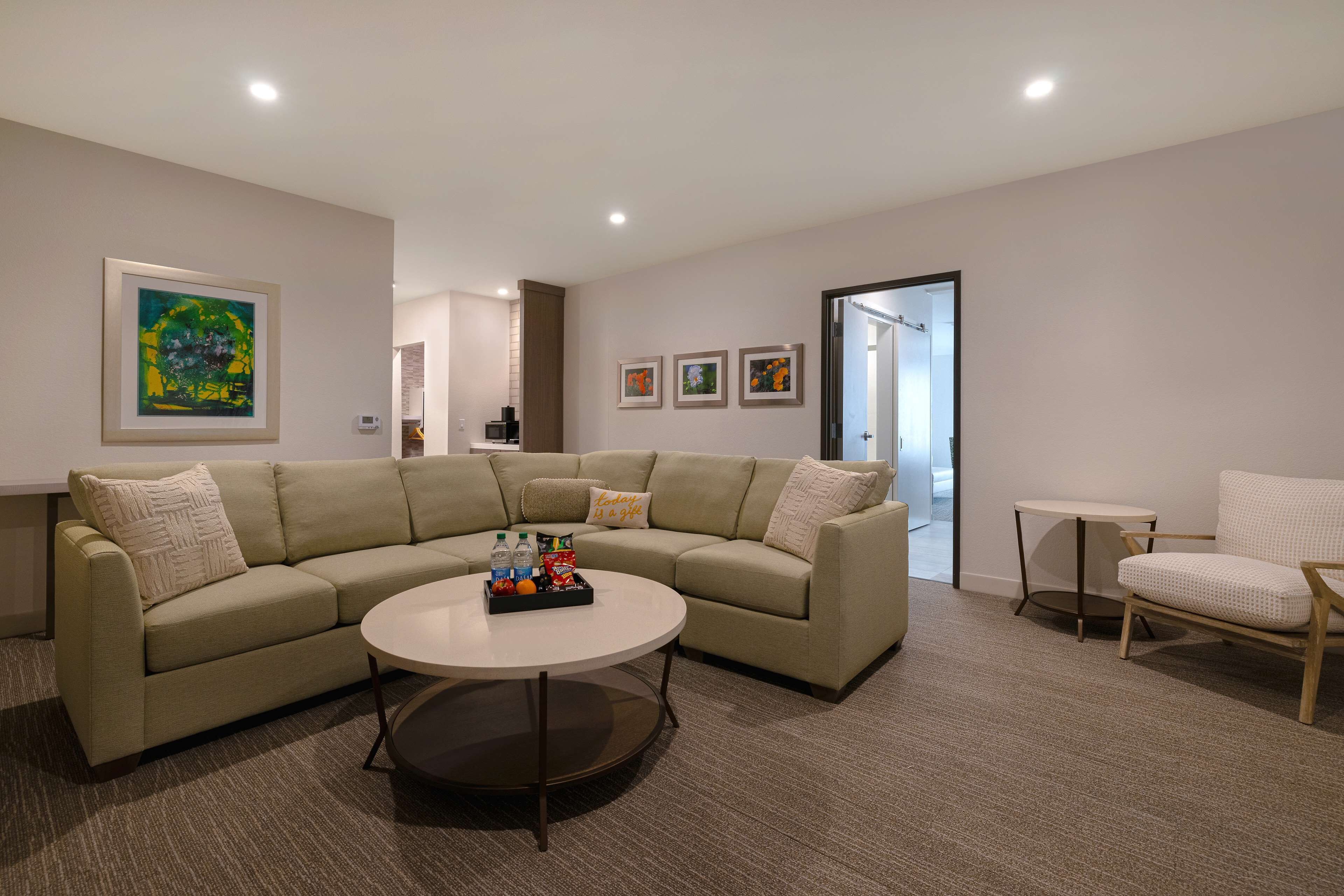 Una sala de estar moderna con un sofá seccional color beige, una mesa de centro redonda y una mesita redonda. Hay cuadros enmarcados en la pared y una puerta que da a otra habitación.