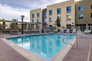 A swimming pool with a metal ladder, surrounded by lounge chairs and a building with multiple windows.