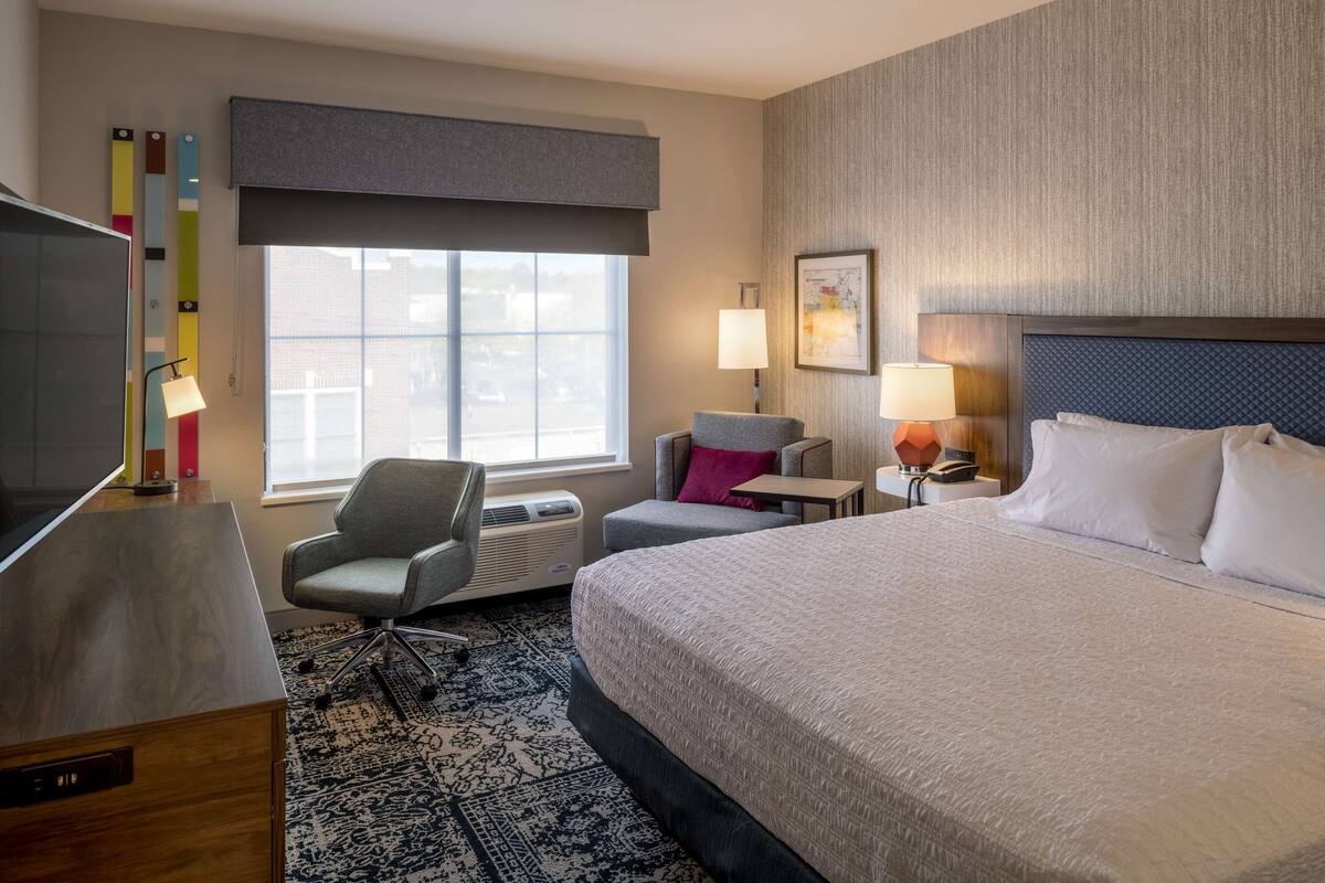 A hotel room with a large bed, a desk, a chair, a small table, a lamp, a map on the wall, and a window with a view of buildings.