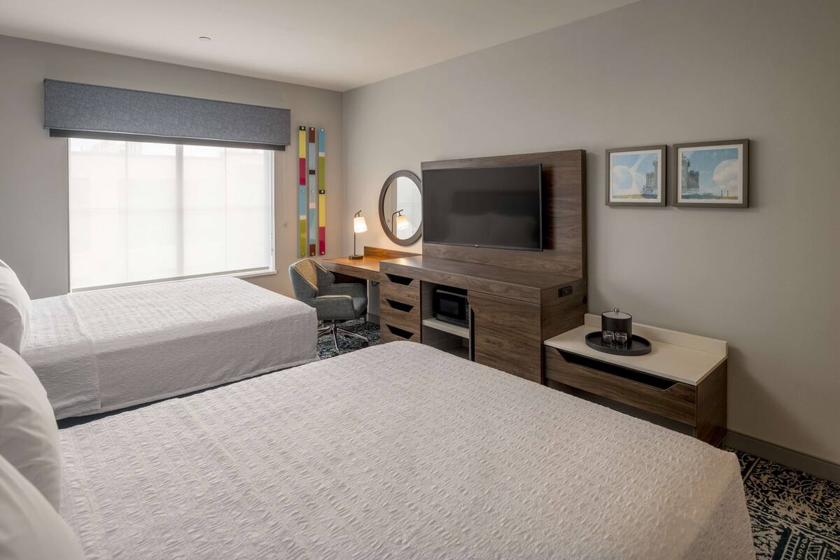 A hotel room with two beds, a desk with a flat-screen TV, a small round mirror, and a side table with a lamp.