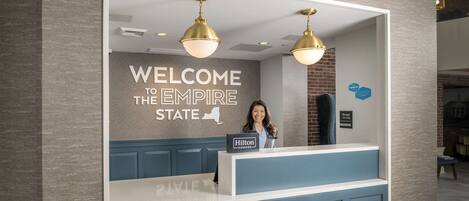 A hotel reception desk with a "Welcome to the Empire State" sign, a "Hilton Honors" sign, and a "Manager On Duty" sign.