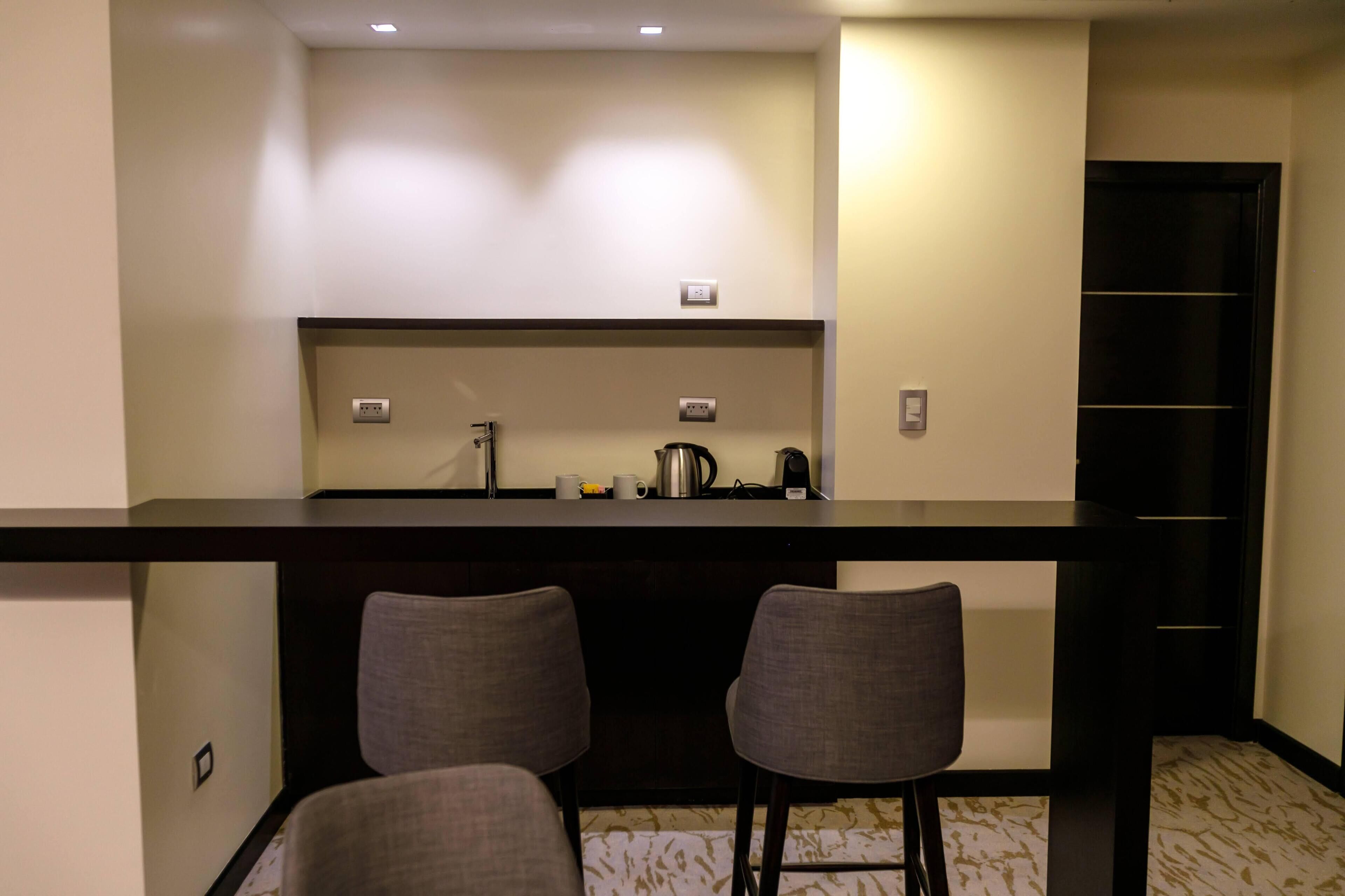A compact kitchenette with a dark countertop, two grey chairs, and a black door.