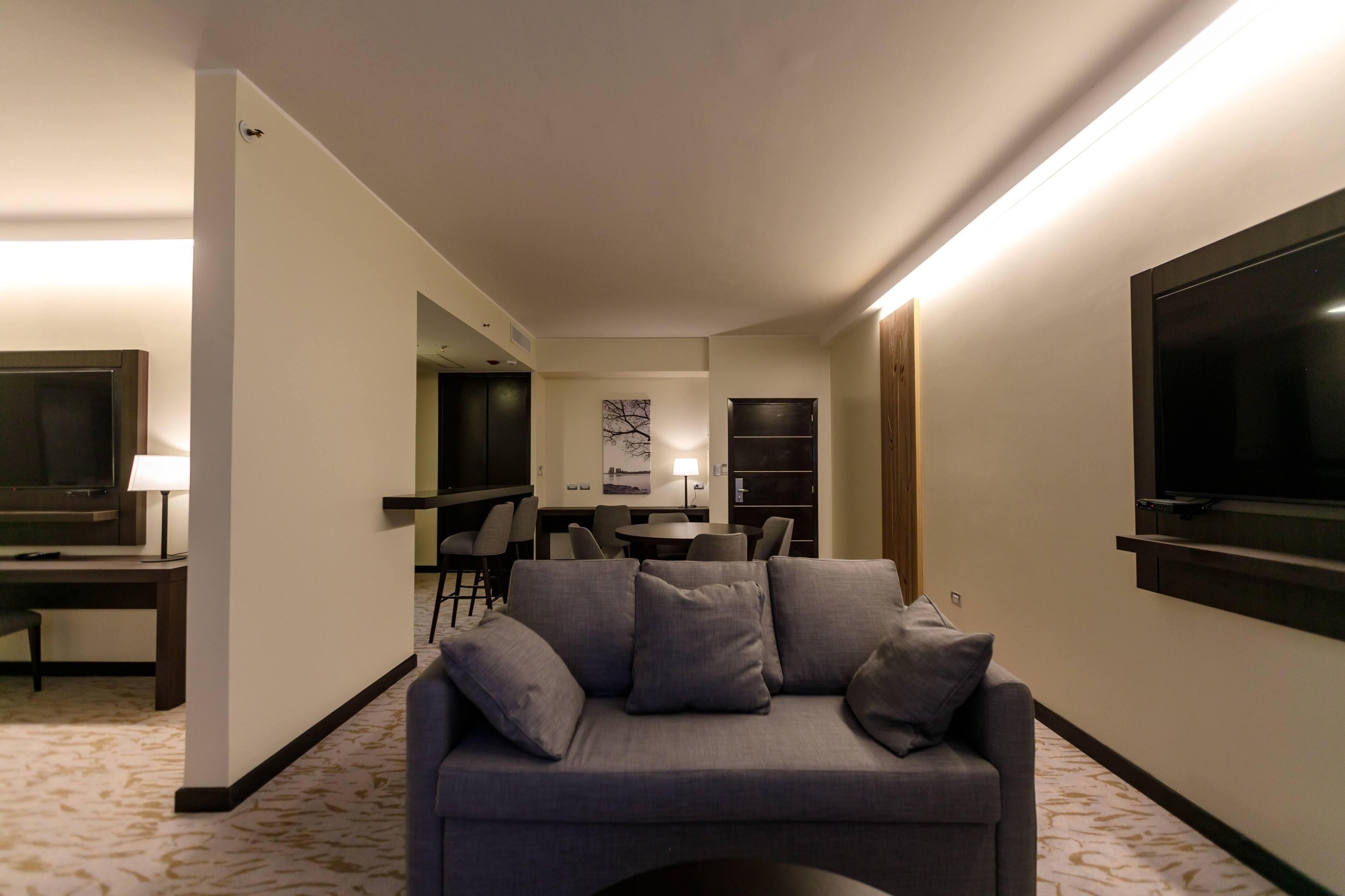 Une chambre d’hôtel moderne avec un canapé gris, un téléviseur à écran plat, un coin repas et une grande fenêtre.