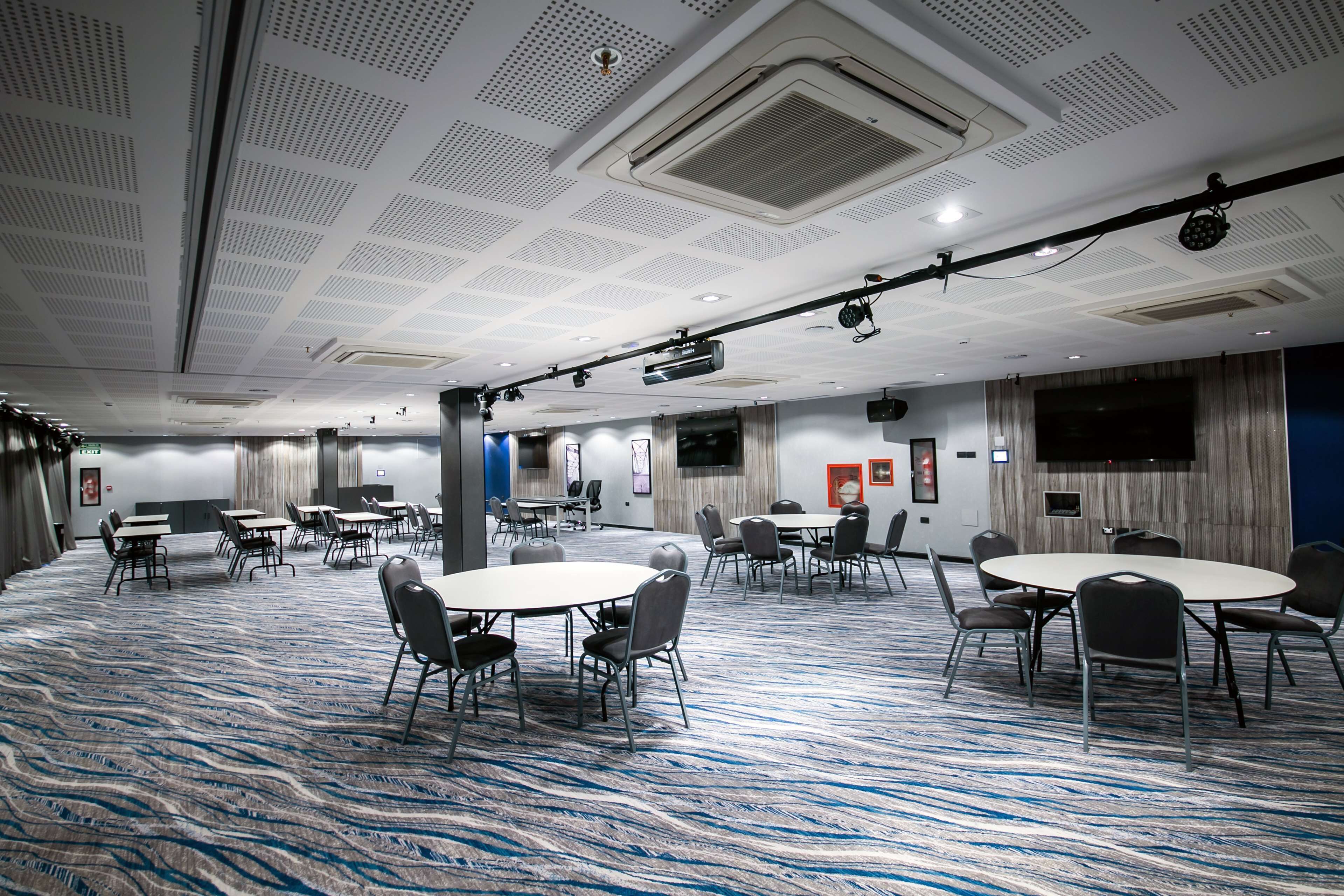 A conference room with multiple tables and chairs, ceiling vents, and a flat-screen TV on the wall.