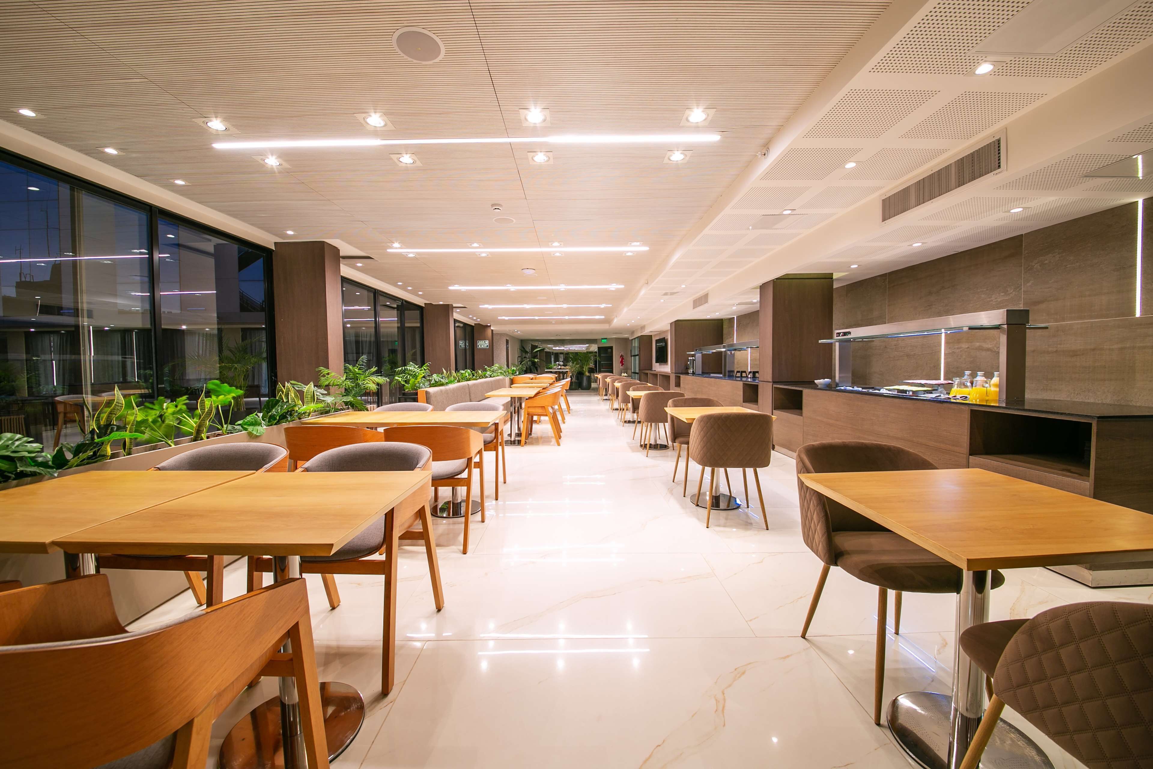 A modern dining area with wooden tables and chairs, a long counter with a kitchen area, and large windows providing natural light.